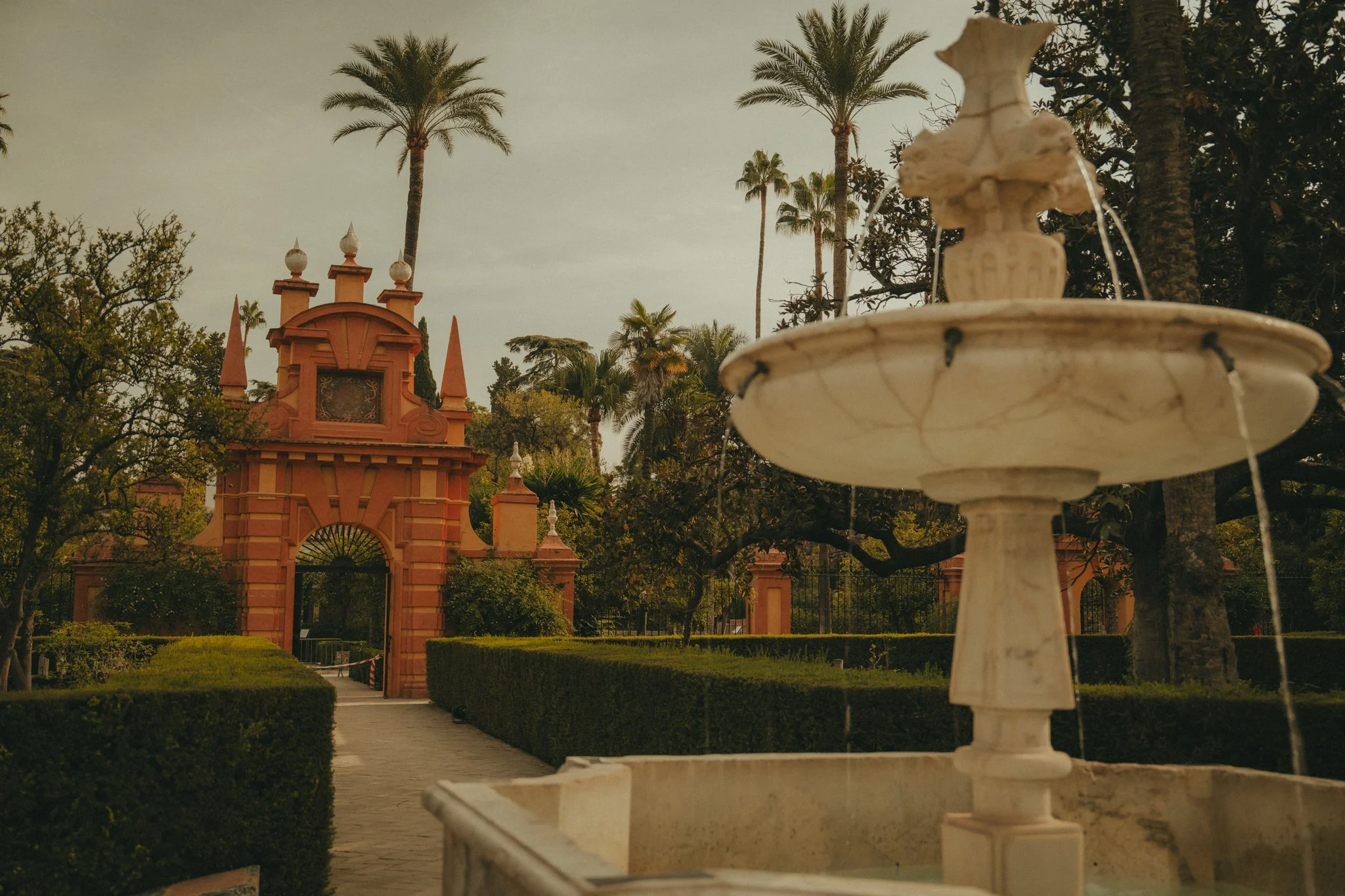 A stone fountain with water flowing into a bowl, situated in front of a historic pink building with towers and spires, surrounded by tall palm trees and lush greenery.