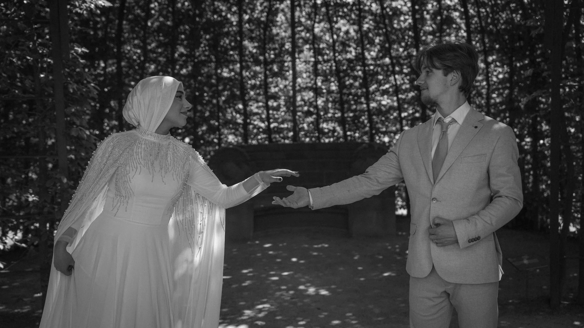 A bride in a white wedding dress and hijab, exchanging vows with a groom in a light-colored suit with a tie, outdoors surrounded by trees.