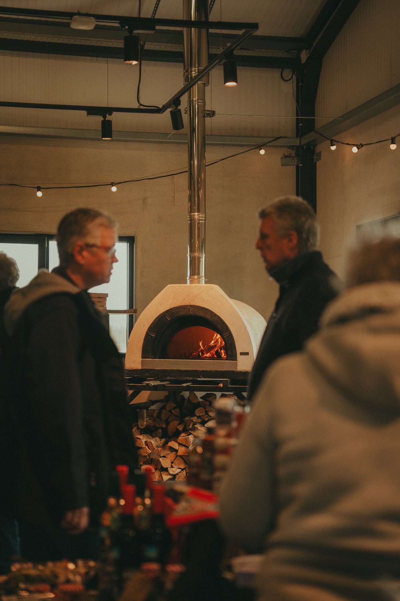 People gather around a fireplace in a cozy indoor setting, with wood logs stacked underneath and string lights hanging from the ceiling.