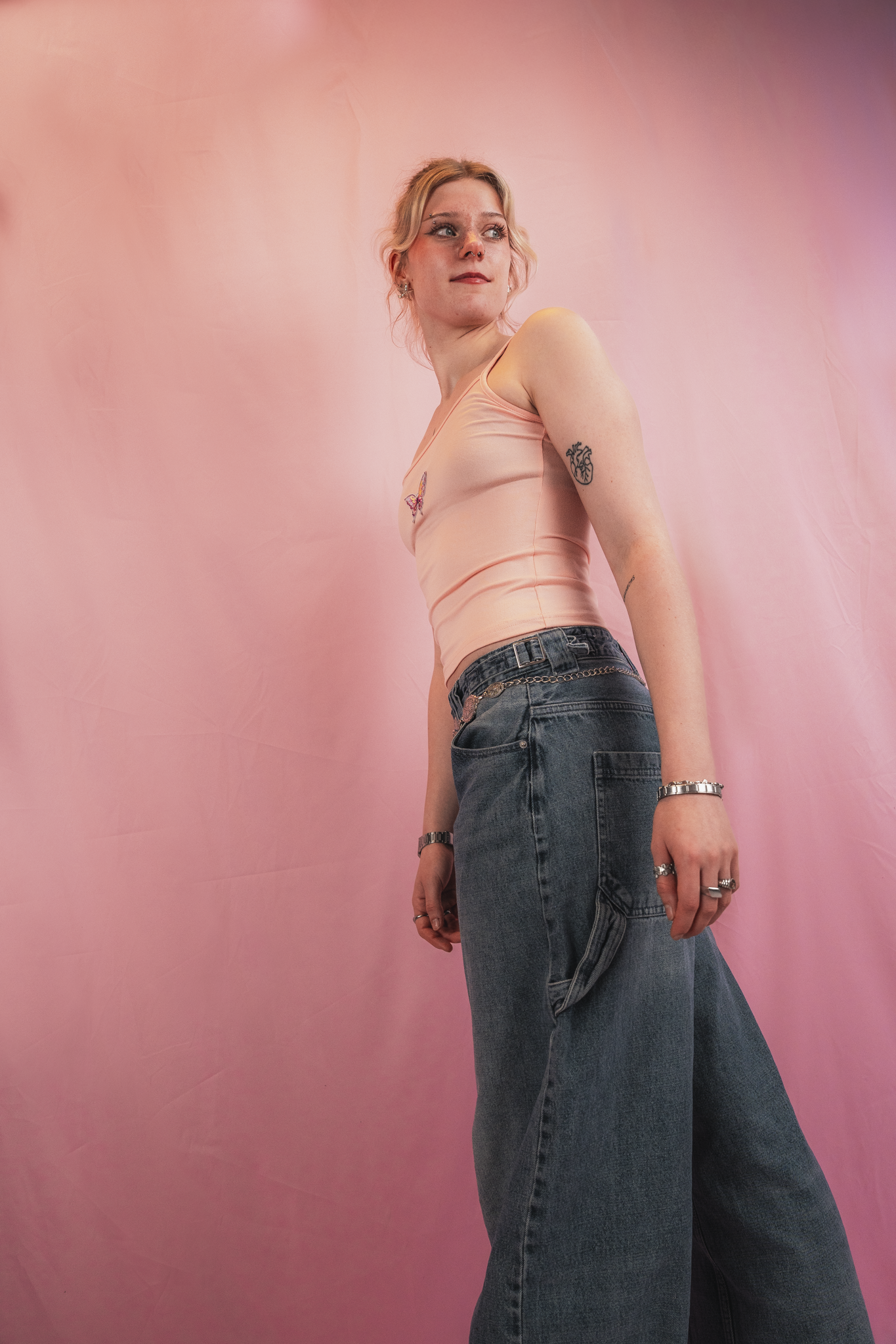 A young woman with blonde hair, wearing a peach tank top with a butterfly embroidery and high-waisted baggy jeans, standing against a pink gradient backdrop, looking to her right with a neutral expression.