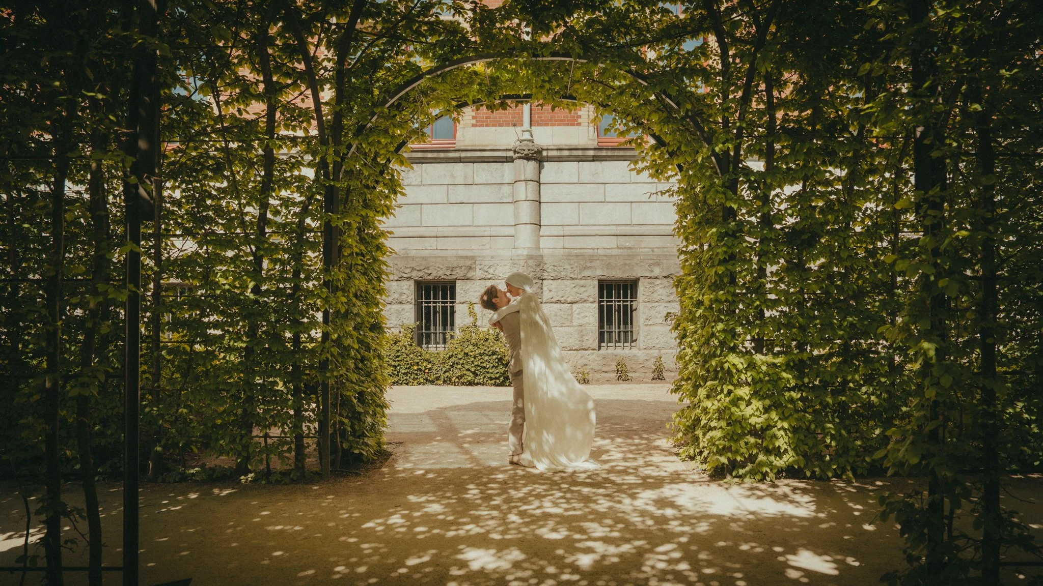 A bride and groom are dancing outdoors under a decorative archway with greenery, in front of a stone and brick building, during daytime.