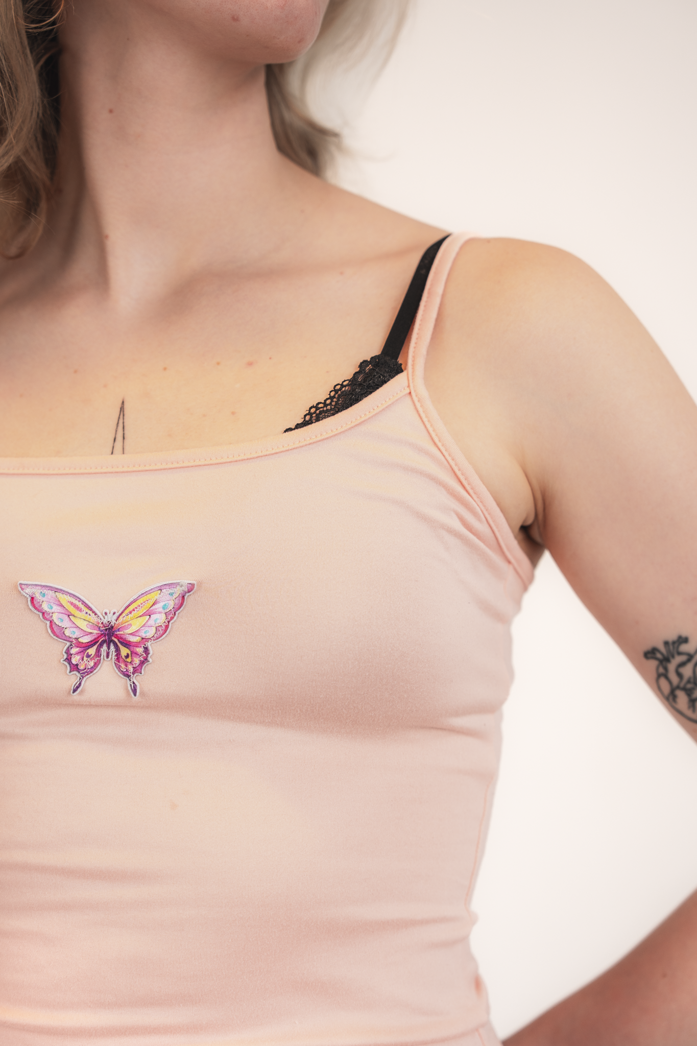 Close-up of a woman's upper chest and shoulder, showing a light pink tank top with a butterfly patch and a black lace undergarment visible at the neckline. She has a tattoo on her forearm.