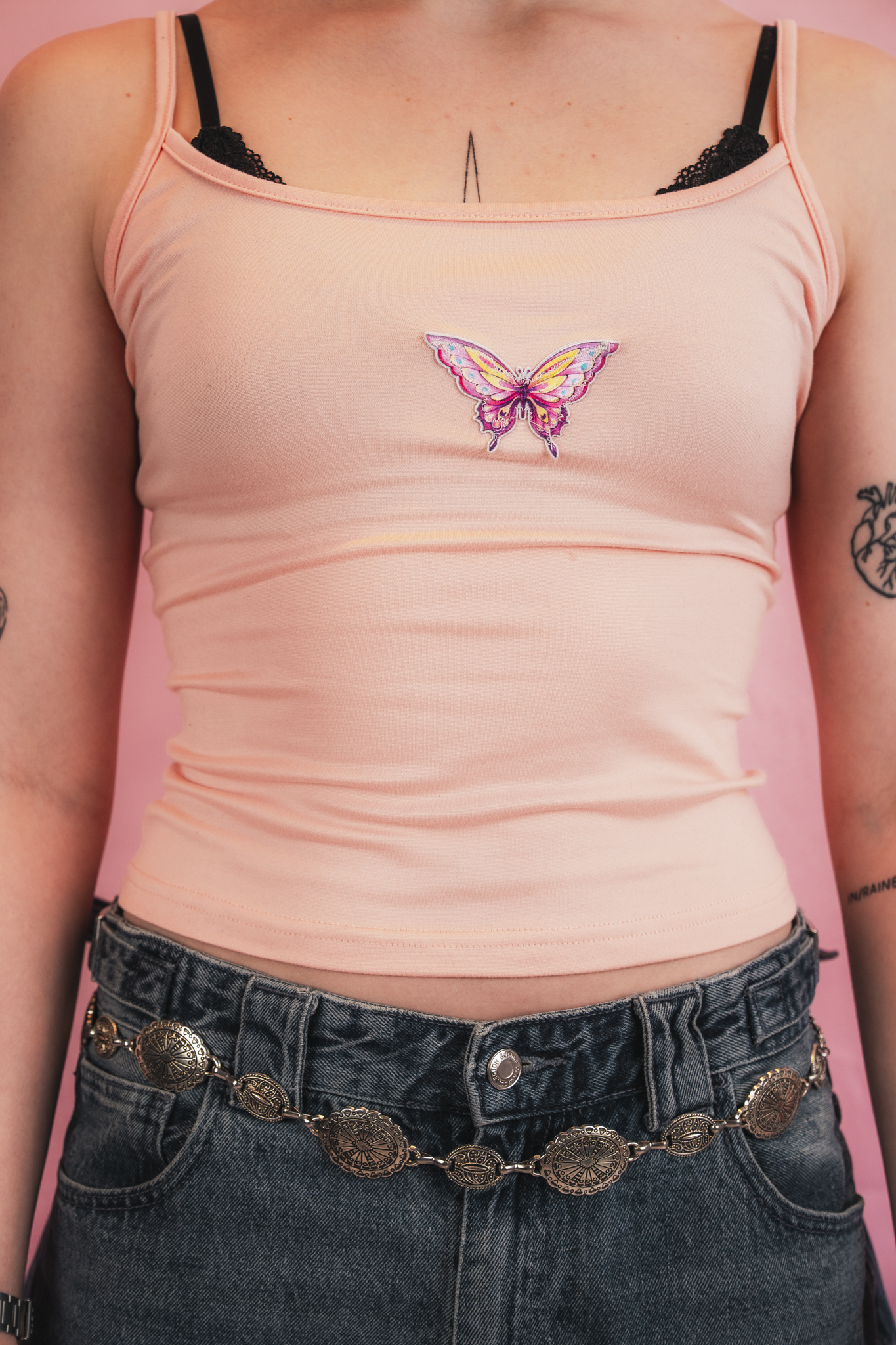 Close-up of a person wearing a blush pink tank top with a butterfly patch on the chest, black lace bra straps visible, and dark high-waisted jeans with a decorative chained belt.