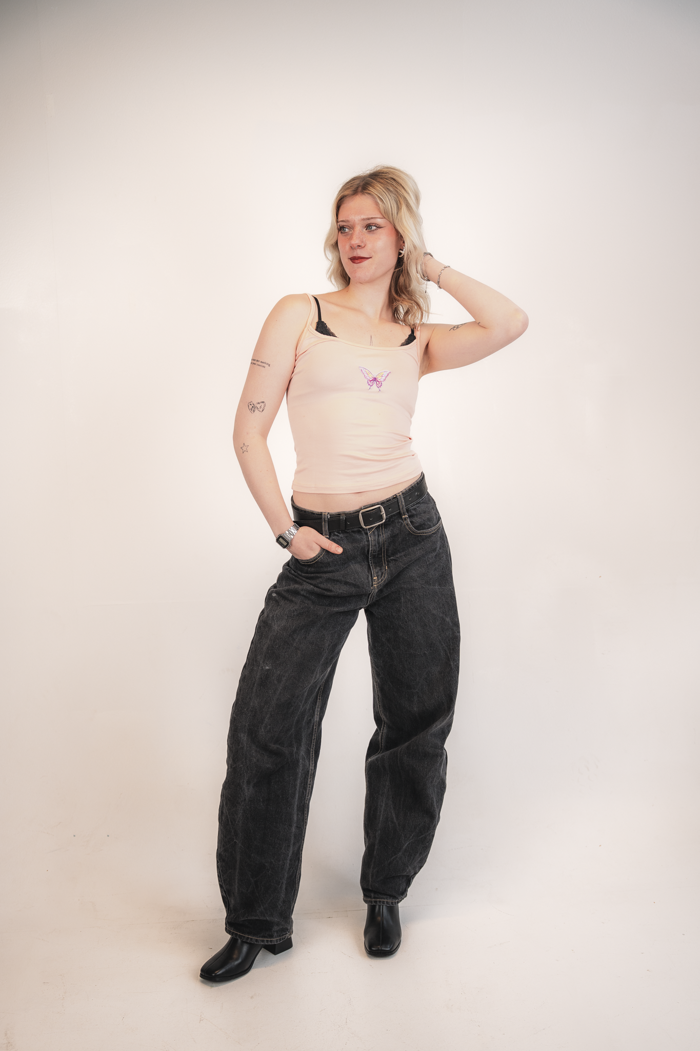 A young woman with blonde hair, wearing a pink tank top with a butterfly embroidery, black high-waisted jeans, and black boots, posing against a plain white wall with one hand behind her head.