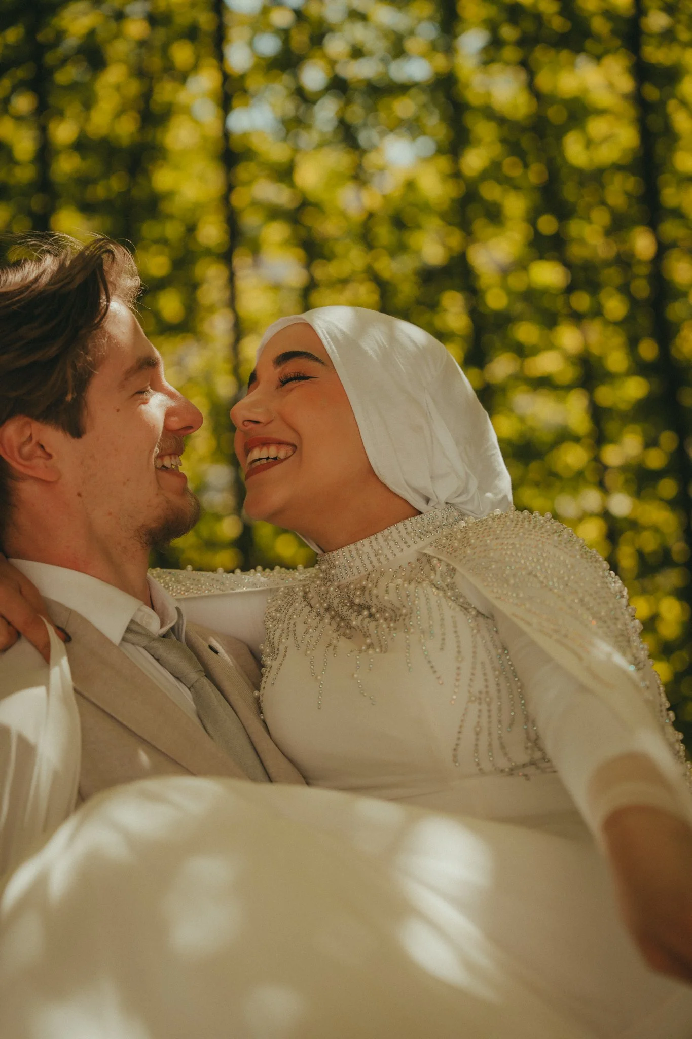 A joyful bride and groom smiling and looking into each other's eyes at their wedding outdoors, with sunlight filtering through the trees in the background.