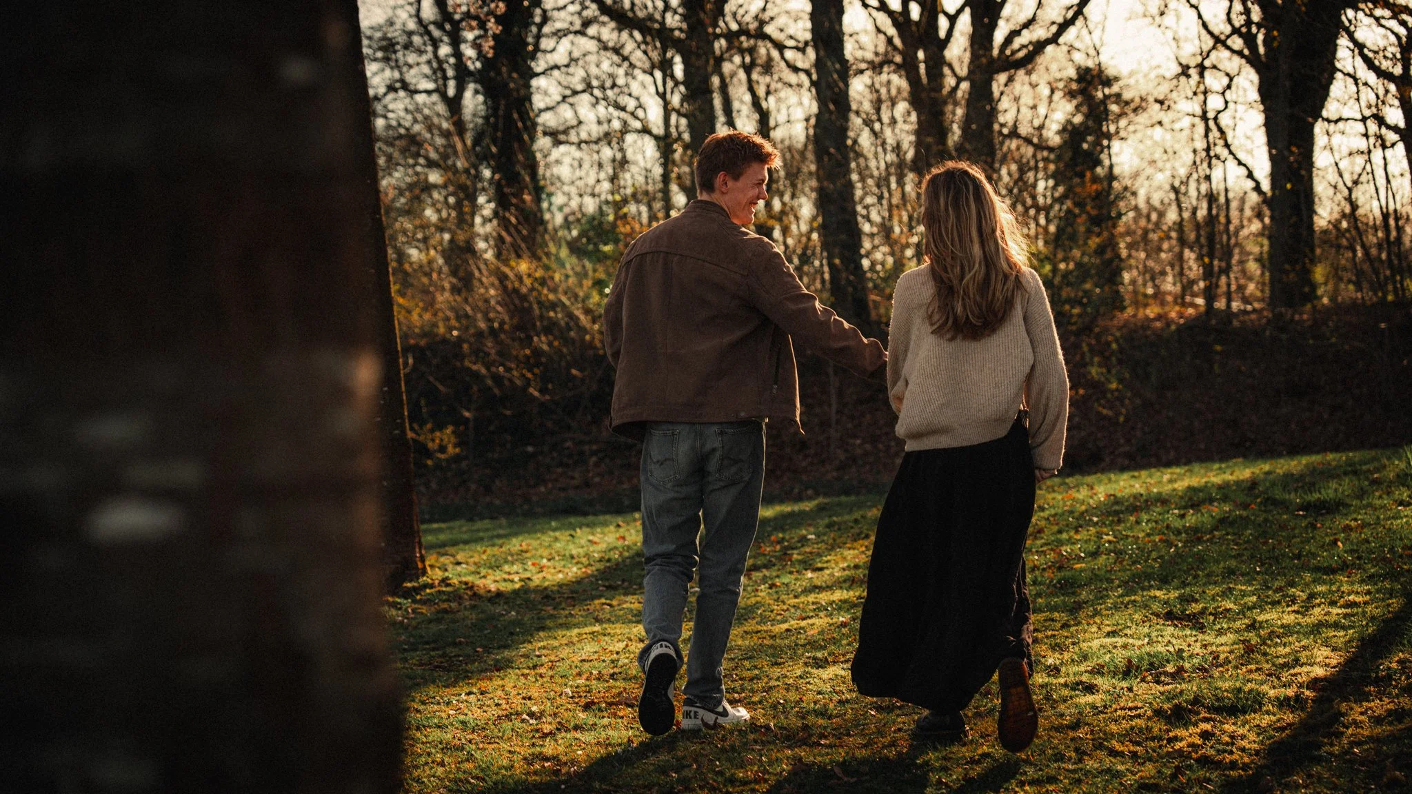 A young man and woman standing in a park during sunset, smiling and holding hands, surrounded by trees with orange and yellow leaves.