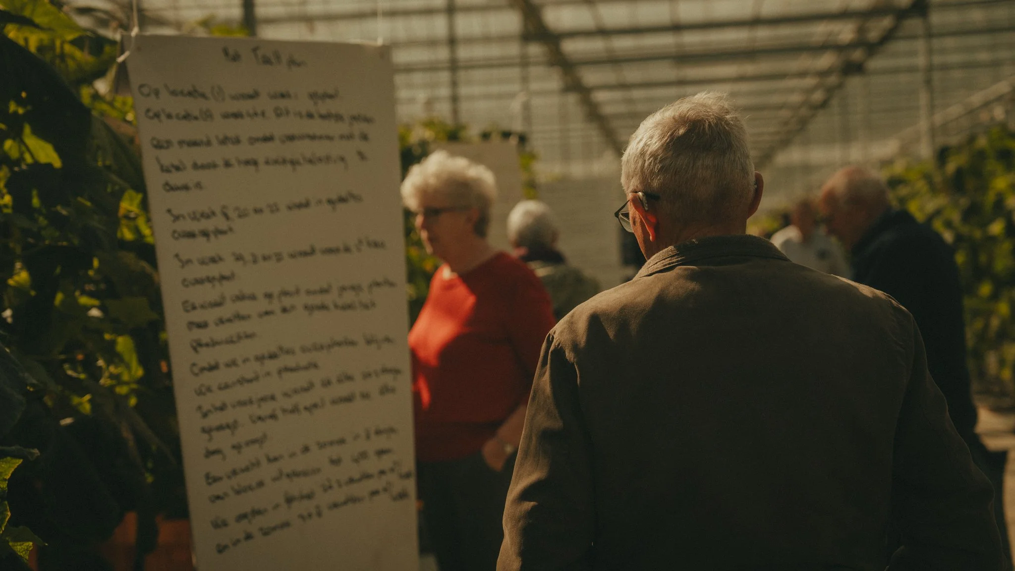 People listening to a speaker in a greenhouse with sunlight and tall plants.