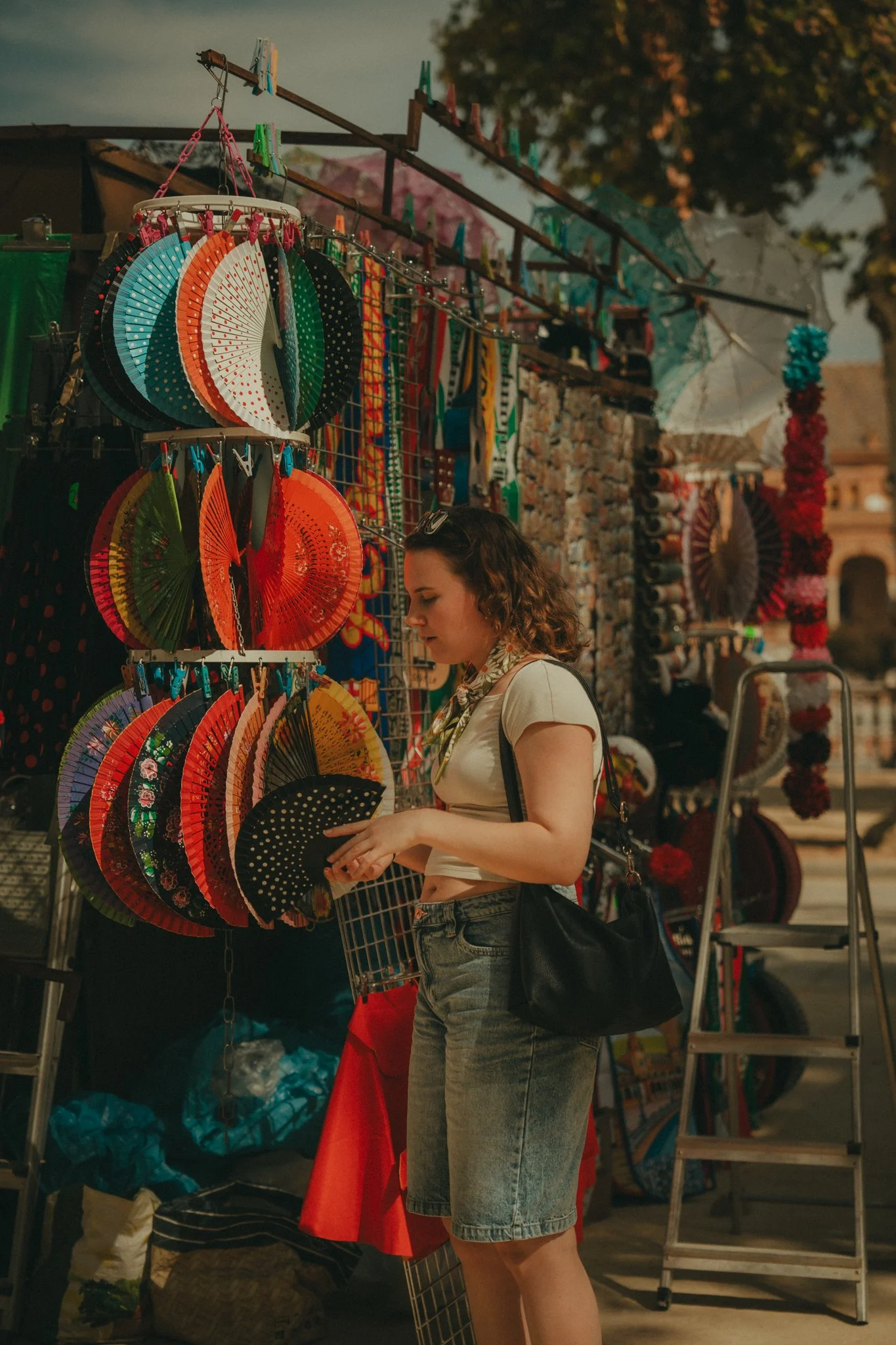 A woman shopping at a street market stall with colorful paper fans and umbrellas displayed. She is wearing a beige t-shirt, denim skirt, and has a black shoulder bag.