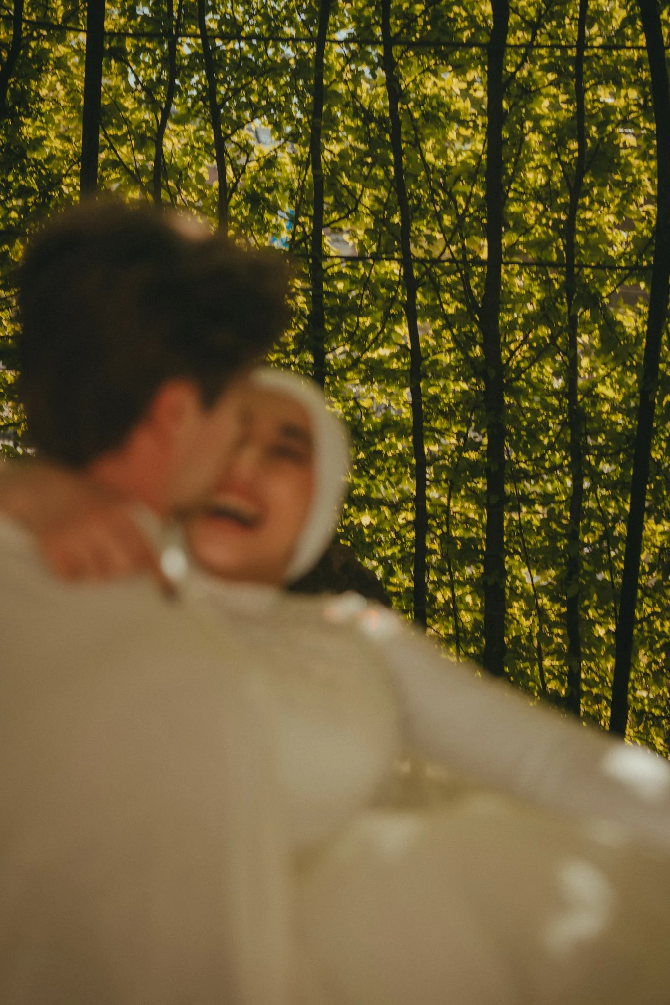 A blurred close-up of two people holding each other, with trees featuring green leaves in the background.