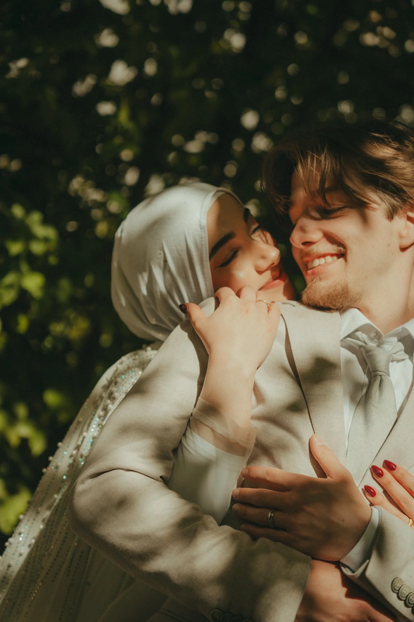 A young couple smiling and hugging outdoors during daylight, with a background of blurred green trees.