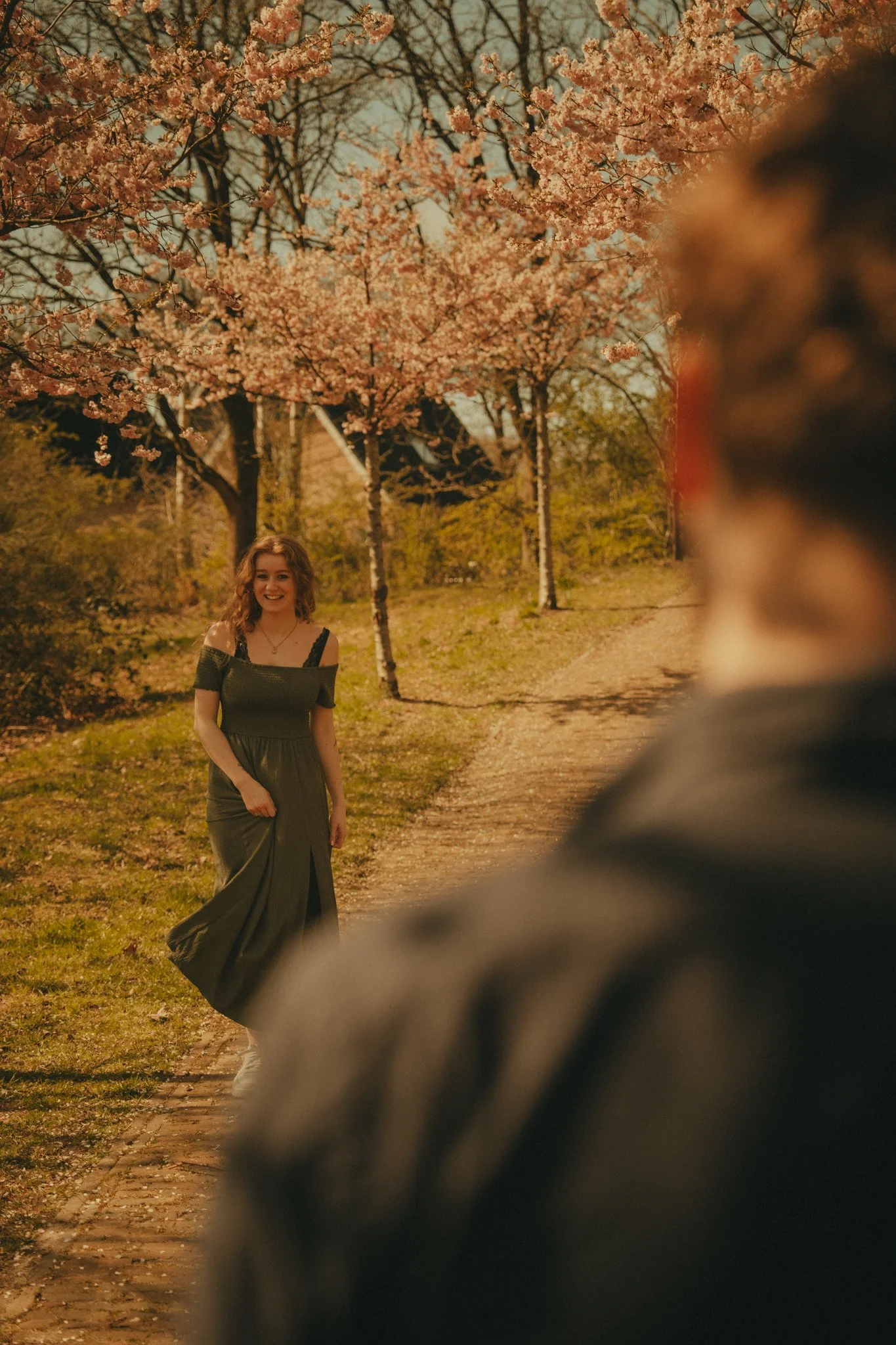 A young woman in a long green dress smiling and walking along a path in a park with blooming pink cherry blossom trees. A person in the foreground is blurred, partially blocking the view.