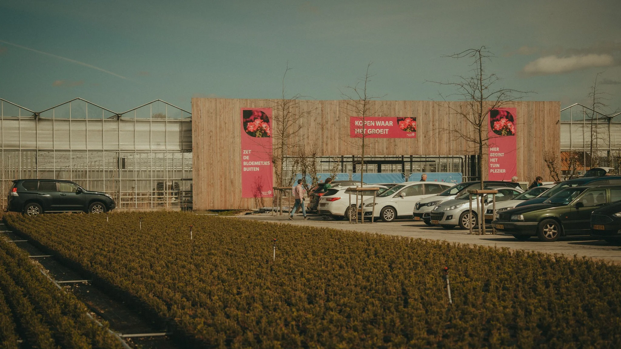 Parking lot with cars in front of a greenhouse building with pink banners displaying flowers, surrounded by leafless trees on a sunny day.