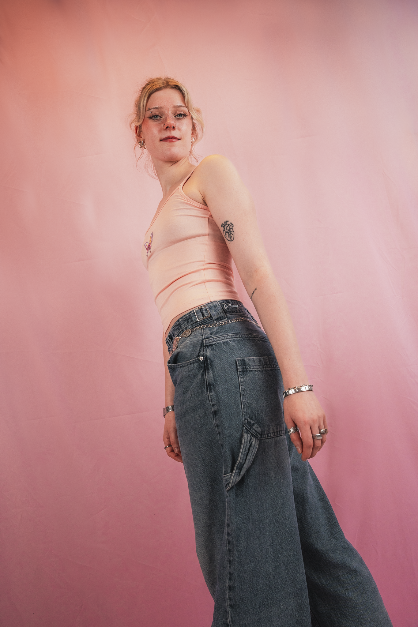 A young woman with blonde hair and tattoos, wearing a peach-colored tank top and high-waisted jeans, standing against a pink background.