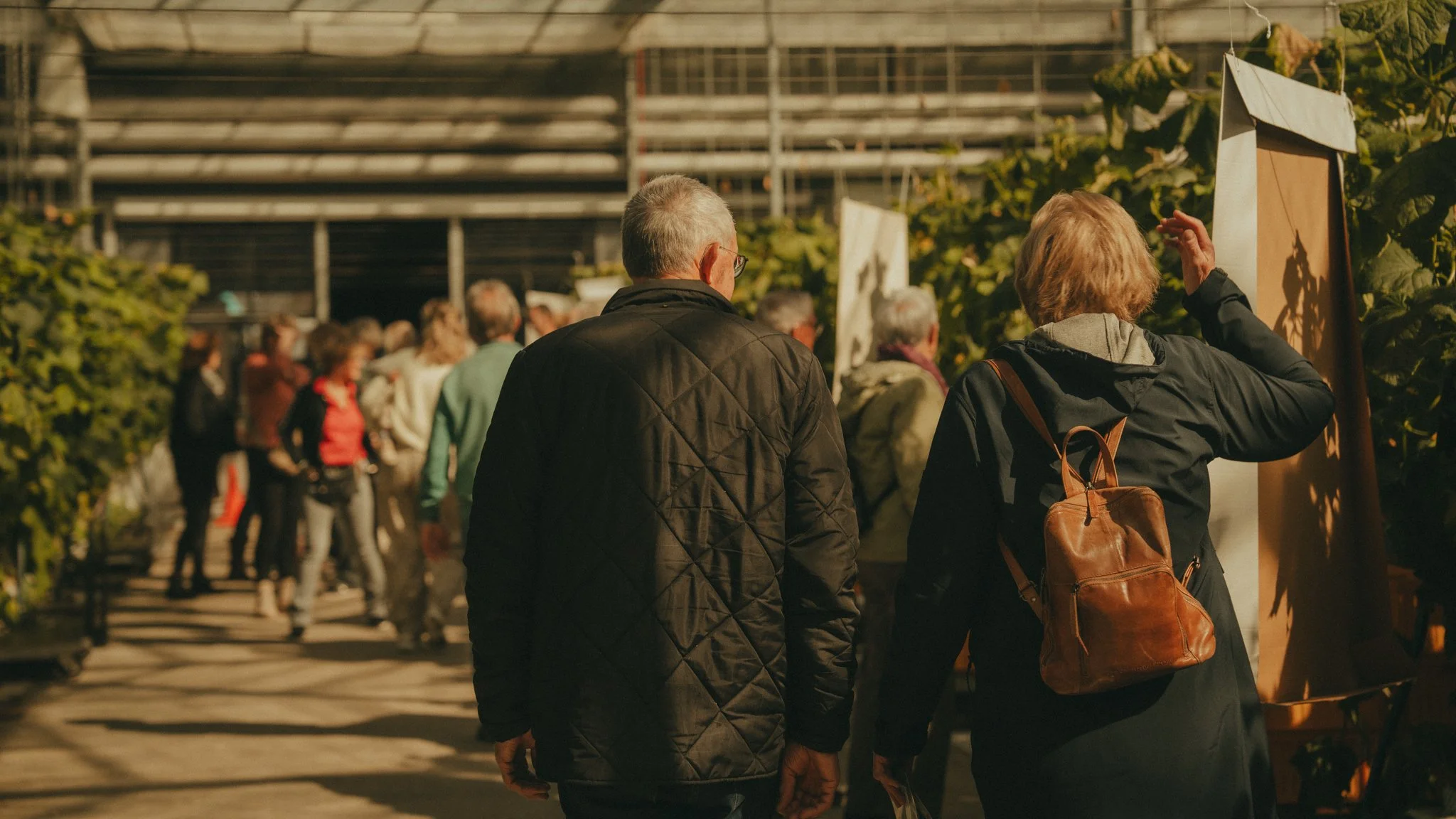 Two women walking through a garden or greenhouse area, with one woman gesturing towards a display or artwork, while other people gather nearby.