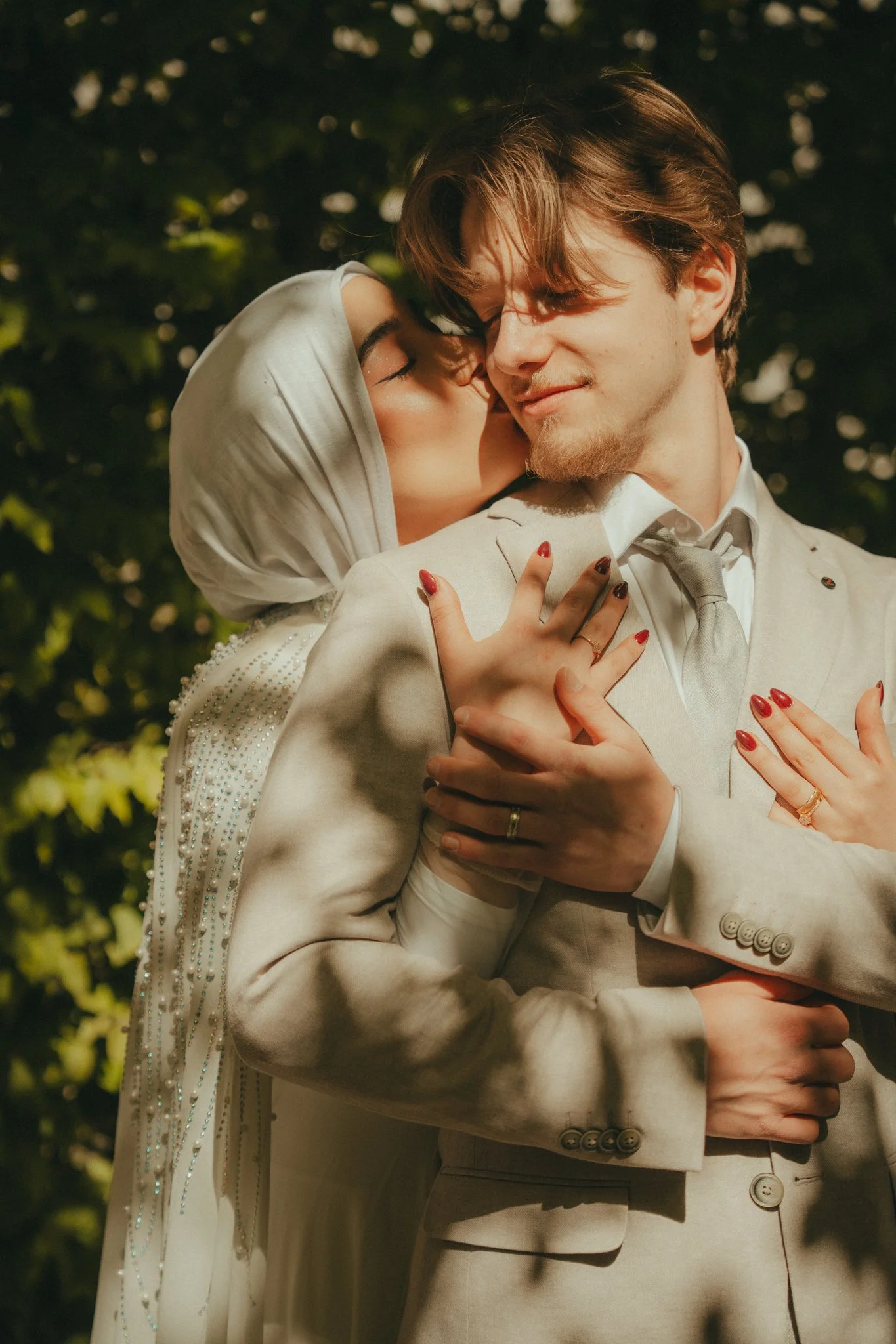 A couple, dressed in formal wedding attire, sharing a tender moment outdoors at night, with the woman kissing the man's cheek and both embracing.