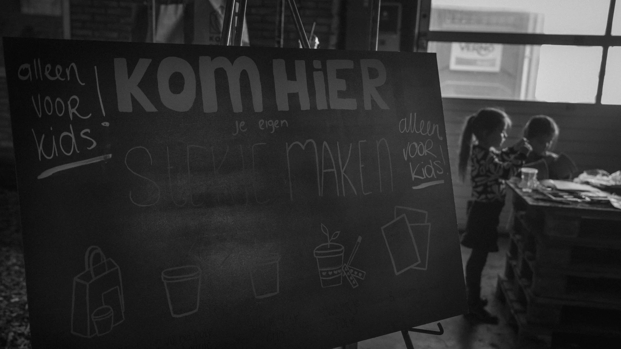 Black and white photo of children in front of a sign that says 'KOMMER voor kids' in a room with large windows.