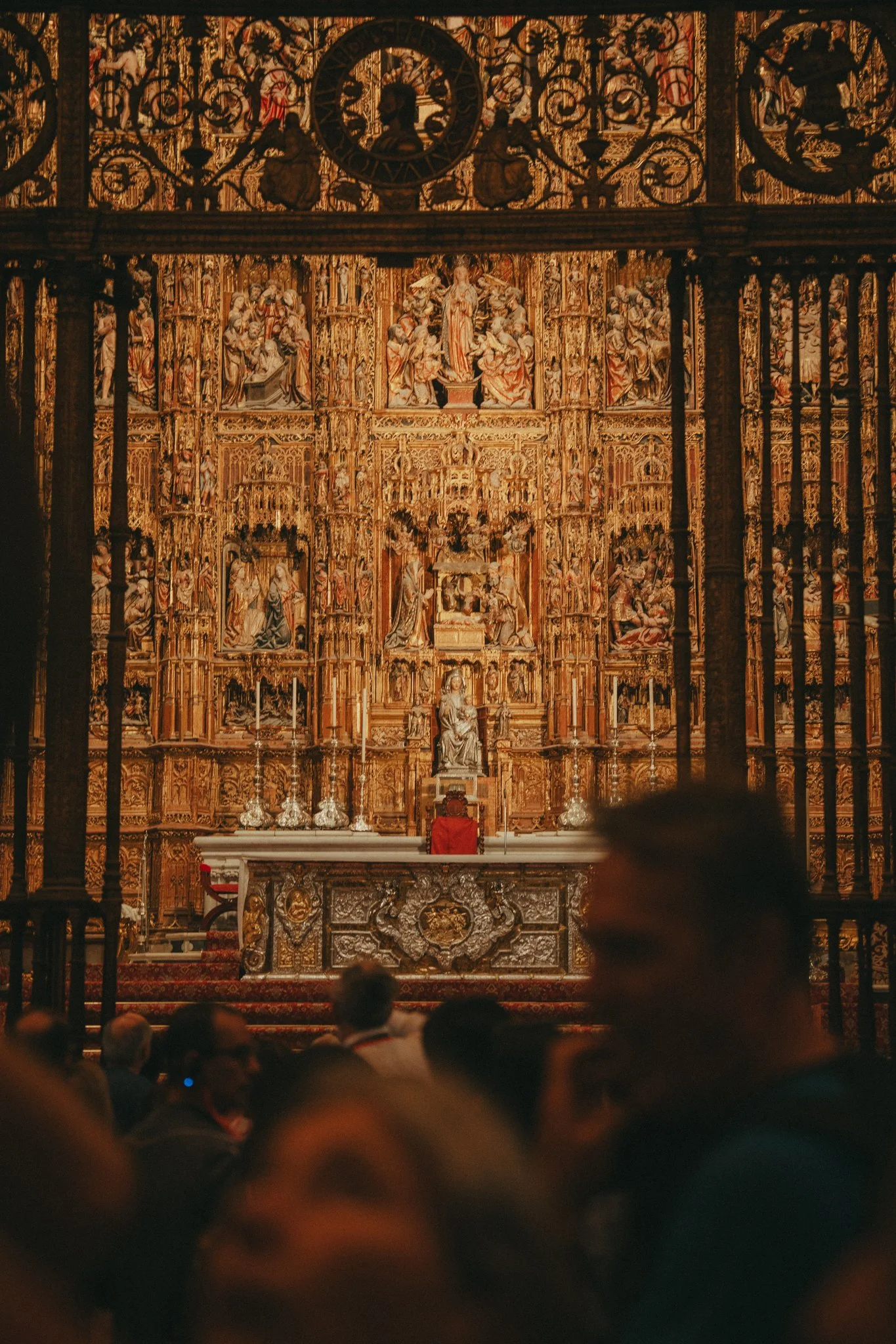 The interior of a church or cathedral with an ornate altar featuring gold detailing, statues, and religious artwork, seen through black iron bars, with an audience seated and facing the altar.