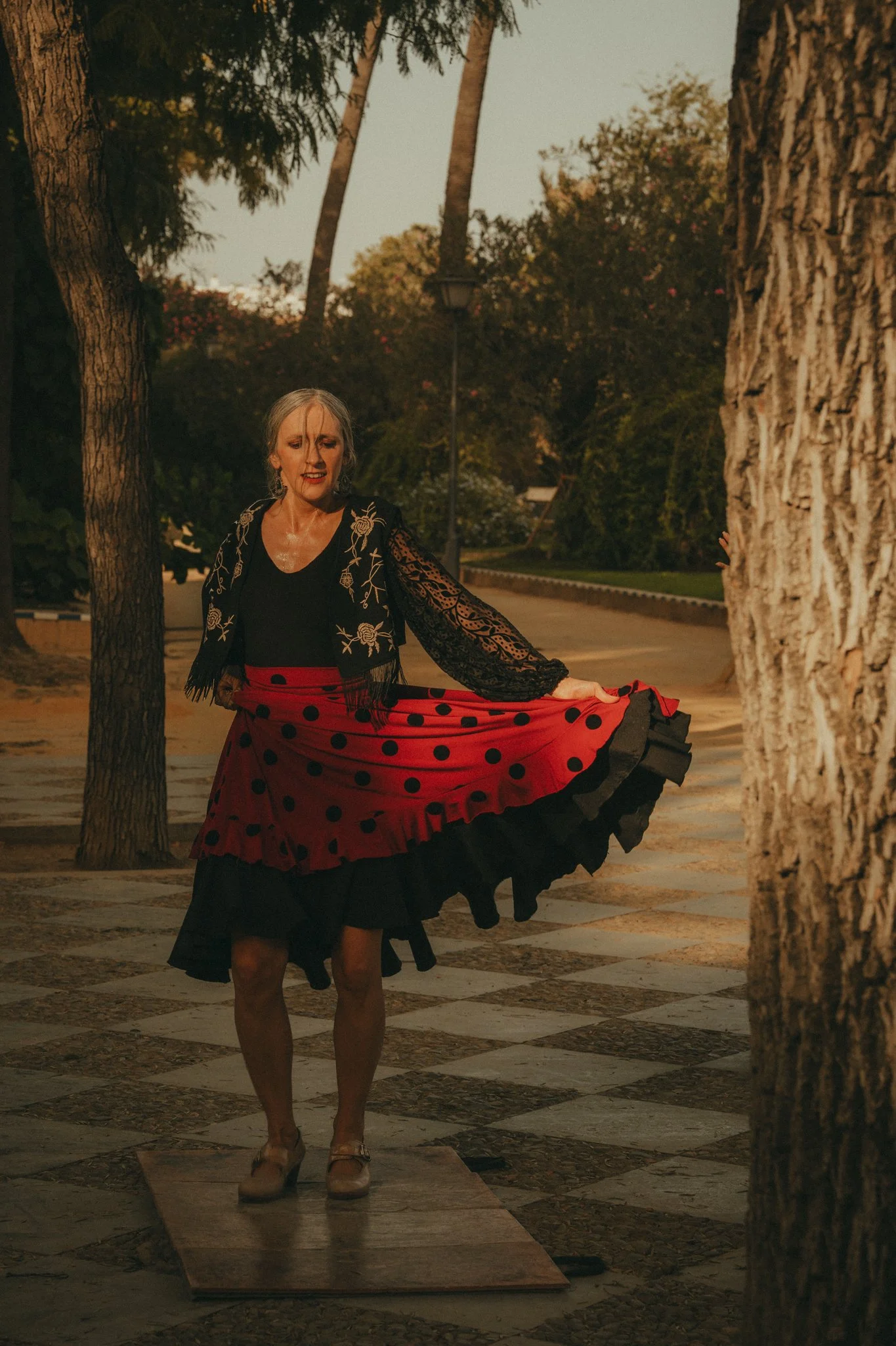 A woman in a flamenco dress dancing outdoors at dusk, surrounded by trees.