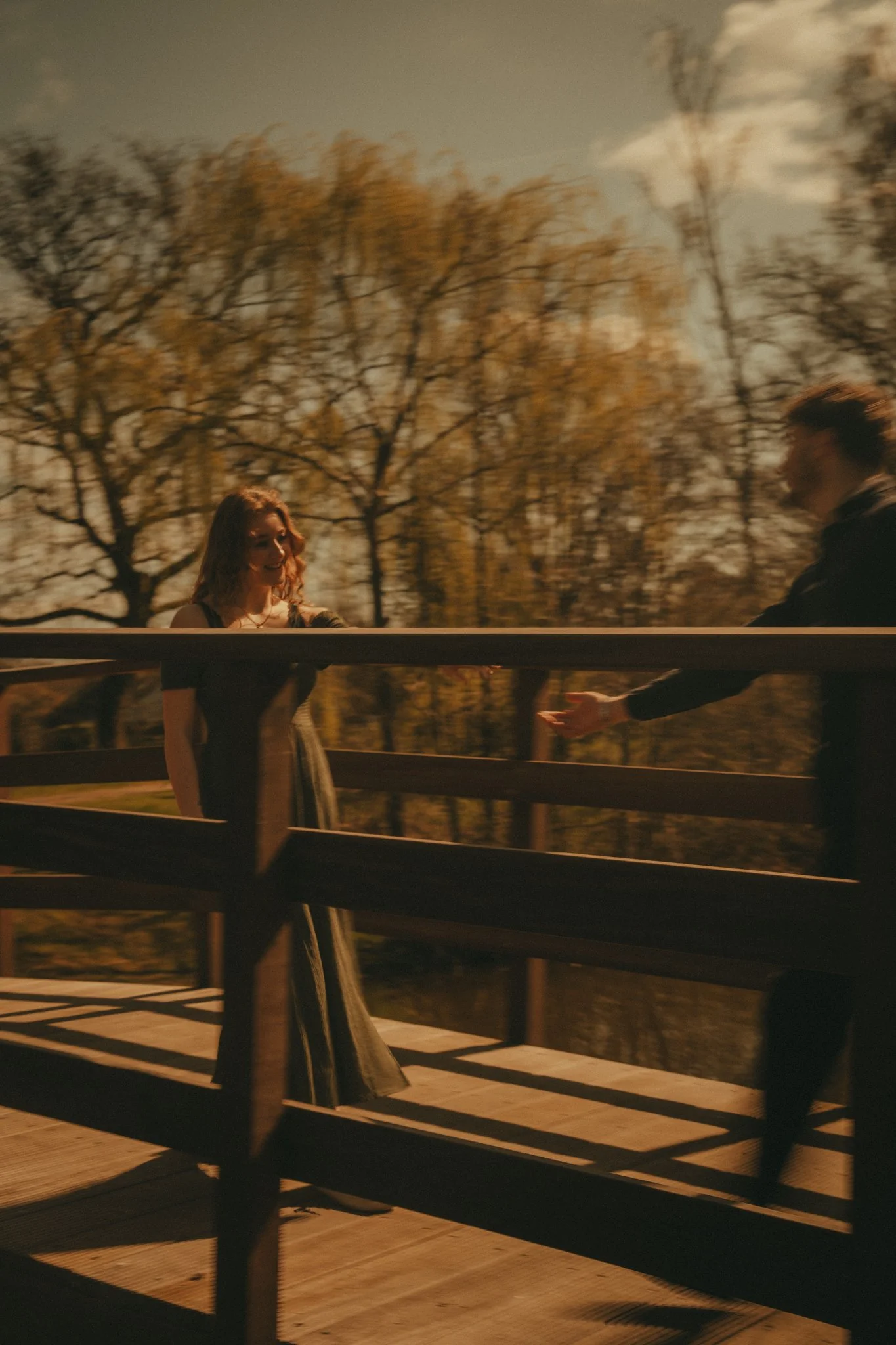 A woman in a black dress smiling and another person extending their hand towards her on a wooden bridge with trees in the background during sunset.