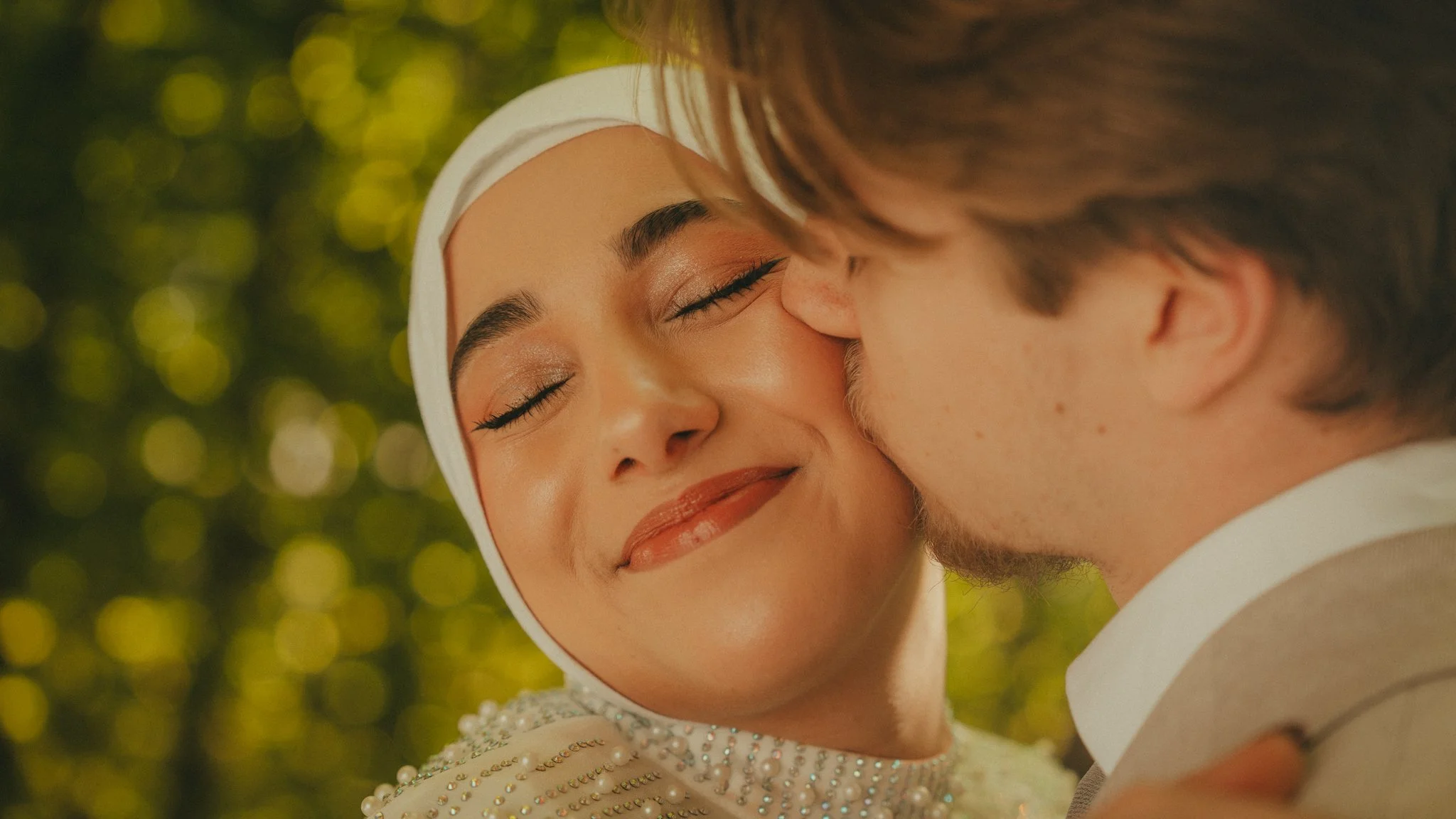 A woman with makeup and a pearl-studded dress is touching her forehead to a man with tousled hair, in a tender moment, with blurred yellow and green foliage in the background.