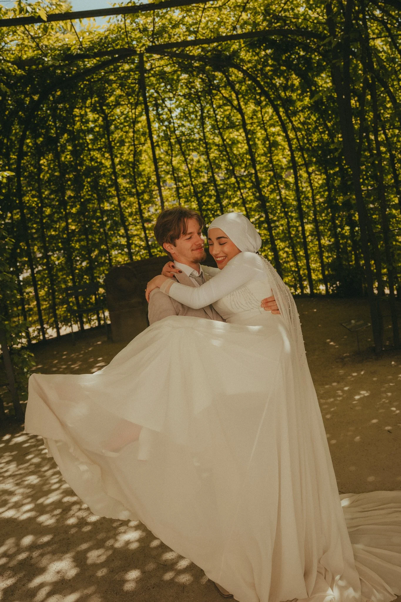 A bride and groom are holding each other and smiling outdoors. The bride is wearing a white wedding dress and headscarf, and the groom is dressed in a light-colored suit. They are standing in front of a black metal fence with green trees and sunlight