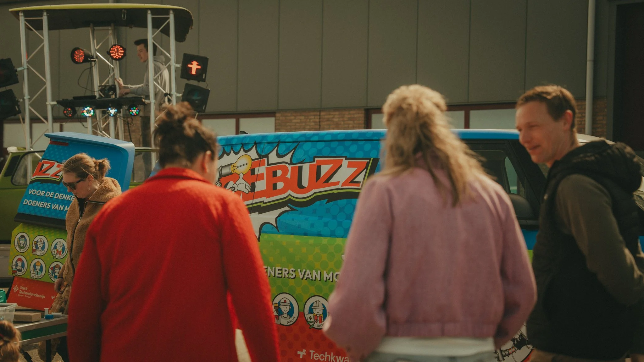 Group of people gathered around a colorful truck with a logo that says 'Buzz', at an event. A DJ is on a raised platform in the background, and there are various posters and signs.