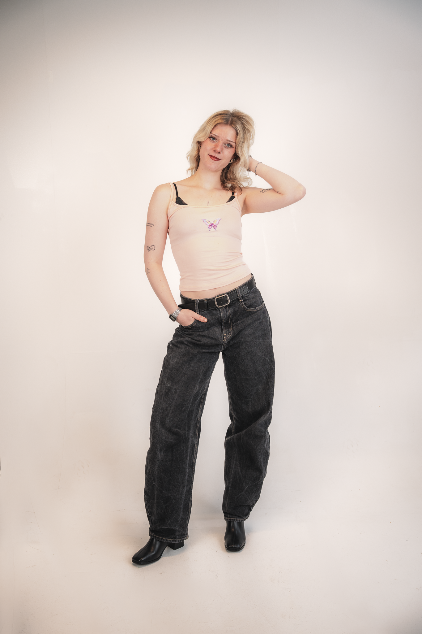 A woman with blonde hair, wearing a light pink tank top with a butterfly design, black lace bra straps visible, black jeans, and black boots, standing against a plain white background with one hand behind her neck and the other in her pocket.