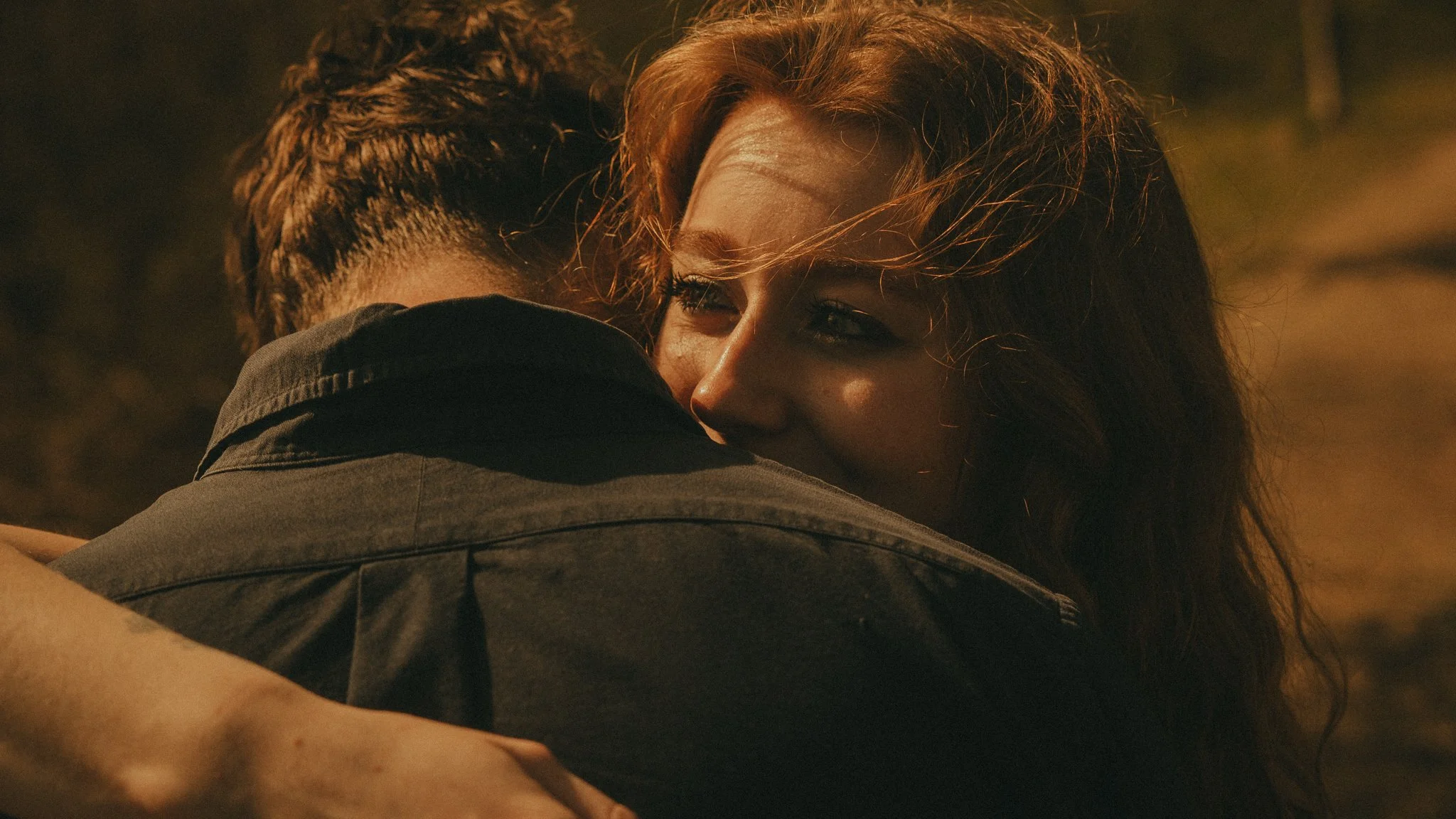 Two people hugging, one with short hair and the other with long red hair, shown from the back and side, outdoors during sunset.