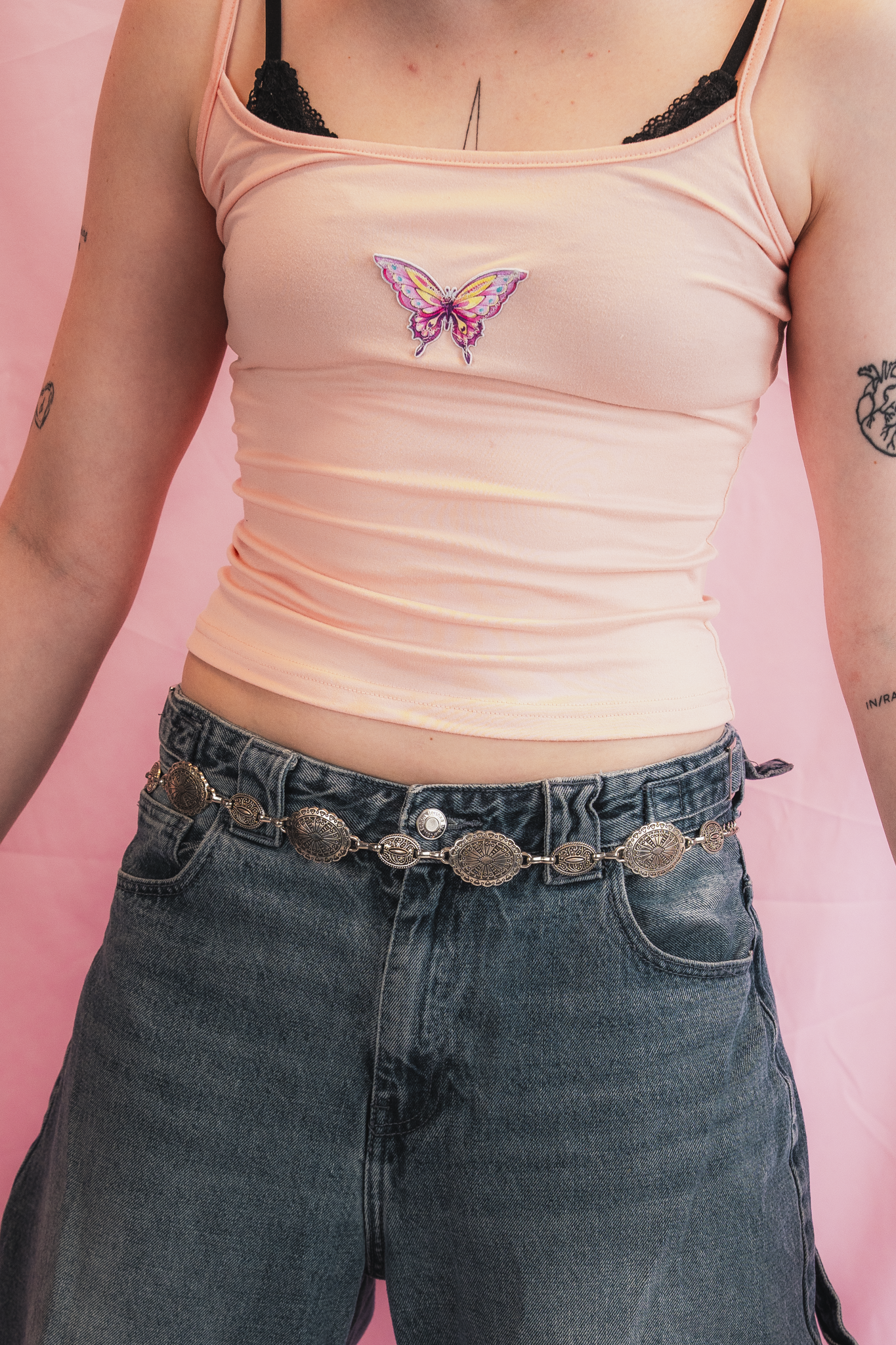 A person wearing a pink tank top with a butterfly patch in the center, black lace bra straps visible, and high-waisted blue jeans with a coin belt, standing against a pink background.