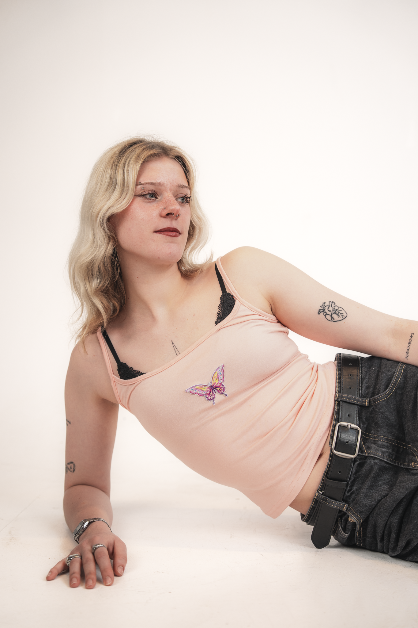 A woman lying on her side against a white background, wearing a pink tank top with a butterfly design, dark jeans, multiple rings, a wristwatch, and tattoos on her arm and shoulder.