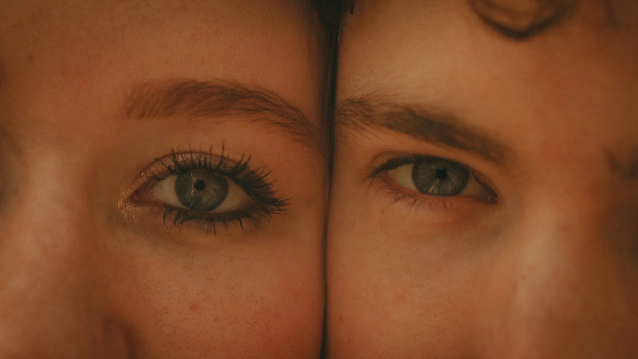 Close-up of two faces with their foreheads touching, showing only their eyes and eyebrows, with similar eye color and makeup.