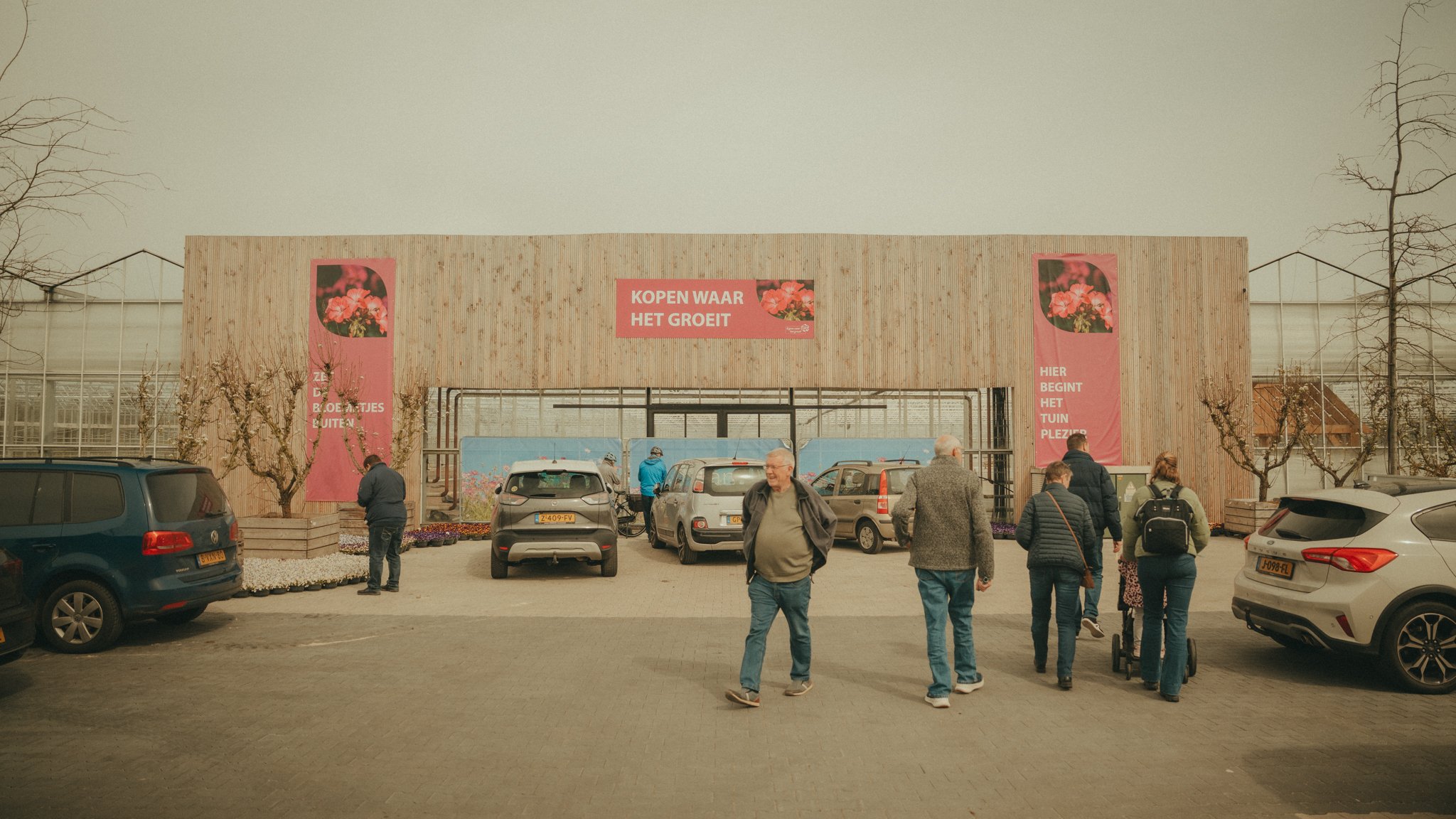 A group of people waiting outside a flower garden shop with pink banners. The building has a wooden exterior and signs in Dutch, including one that says 'KOPEN WAAR HET GROEIT' and other phrases about starting pleasure in the garden.