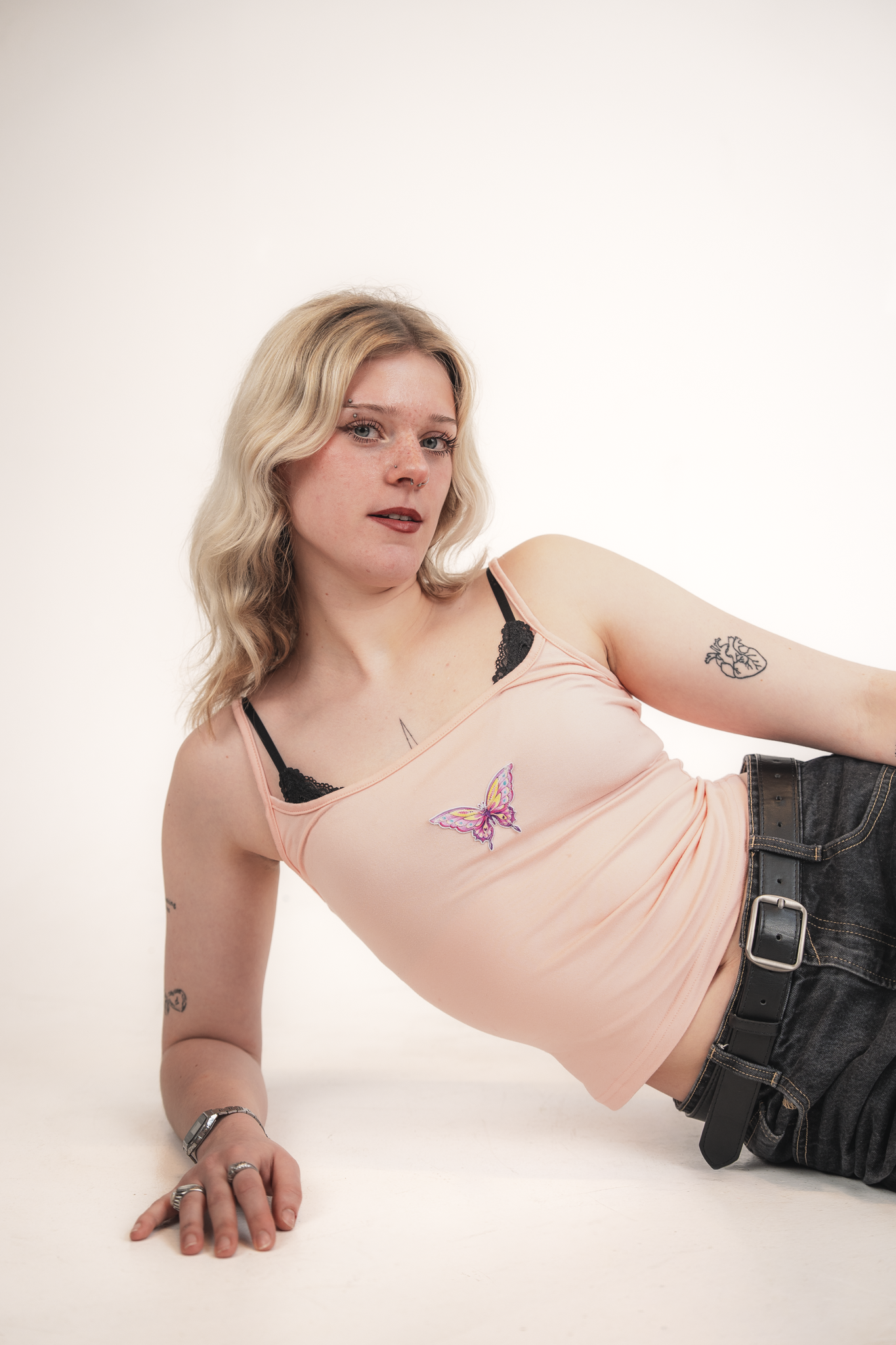 A young woman with blonde wavy hair, facial piercings, and tattoos, lying on her side with her arm extended forward, wearing a peach top with butterfly embroidery and black jeans.