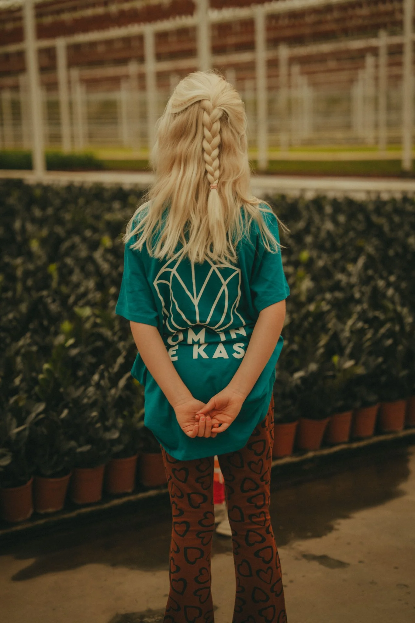 Back view of a person with long, blonde hair styled in a braid, wearing a teal T-shirt with text and dark pants with a heart pattern, standing in front of potted plants and a fence.