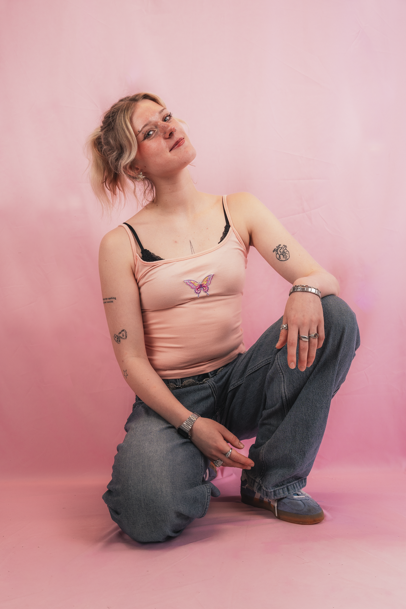 A young woman with short, wavy blonde hair sitting on the floor against a pink backdrop. She is wearing a peach tank top with a butterfly design, gray jeans, and sneakers. She has tattoos on her arms and is wearing jewelry including rings and a watch
