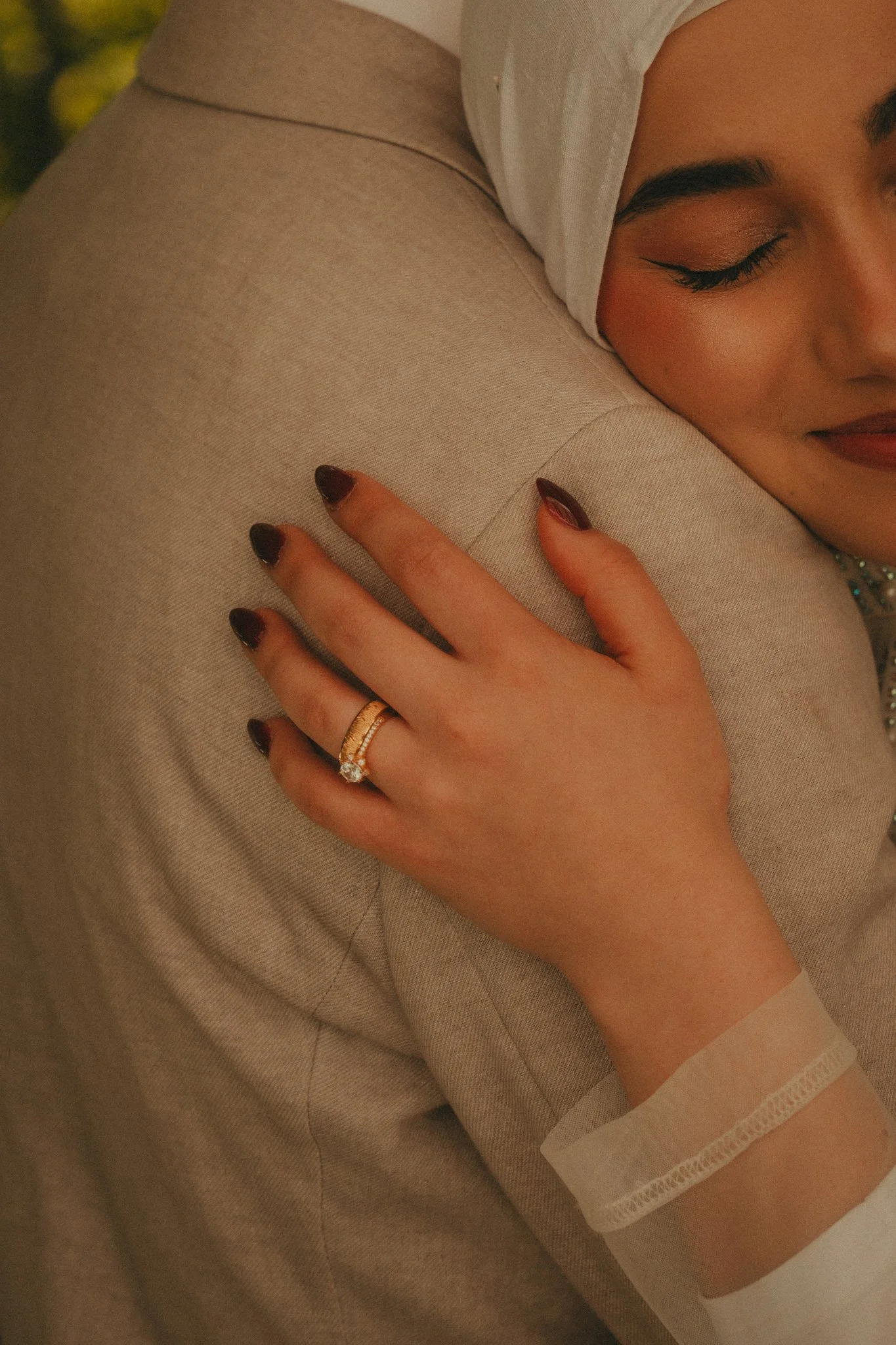 A woman with makeup and ring on her finger is hugging a beige upholstered chair, with her face resting on it and her hand on the chair.