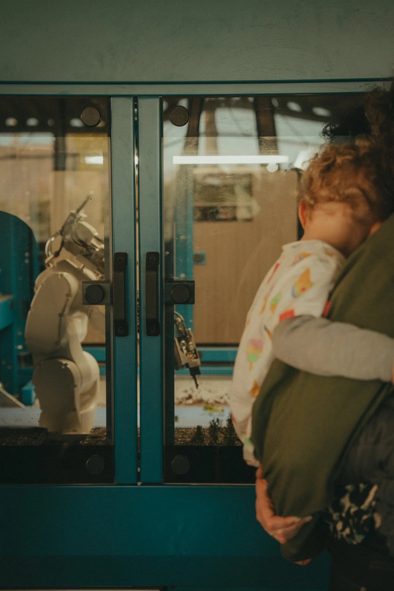 A person with curly hair holding a small child in a green jacket next to a glass door with visible mechanical equipment behind it.