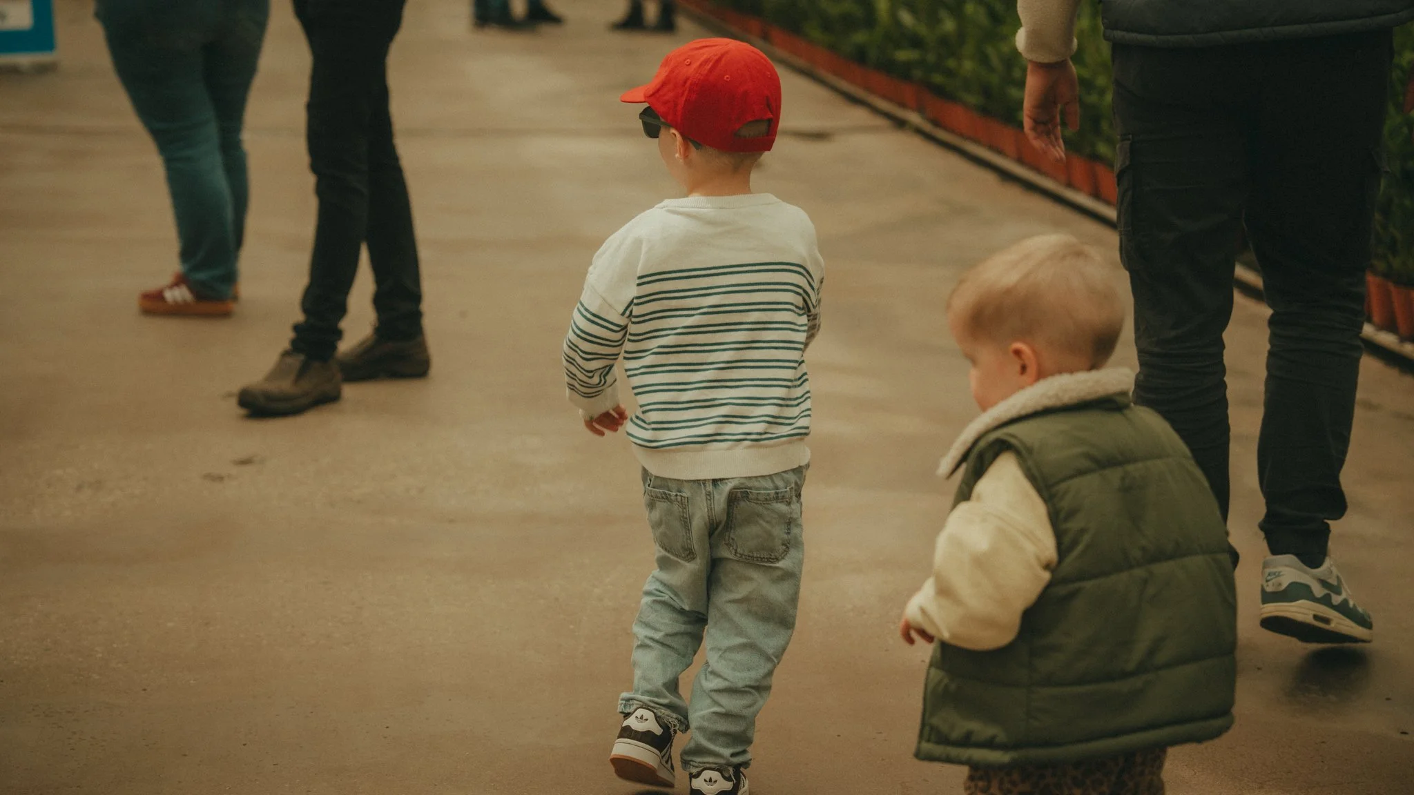 Two young boys walking in a public indoor space, one wearing a red cap and striped shirt, the other with blonde hair in a green jacket.