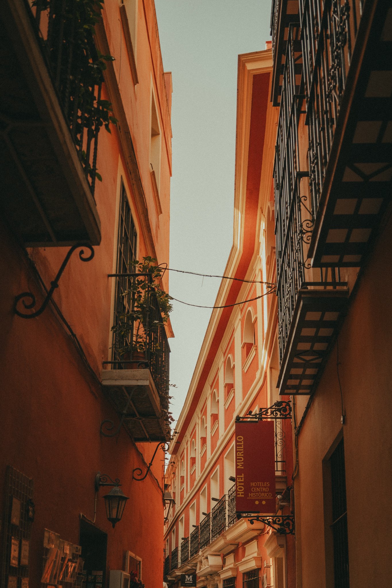 View of a narrow street in a historic district with buildings on both sides, featuring wrought iron balconies and warm-colored facades, under a pale sky.