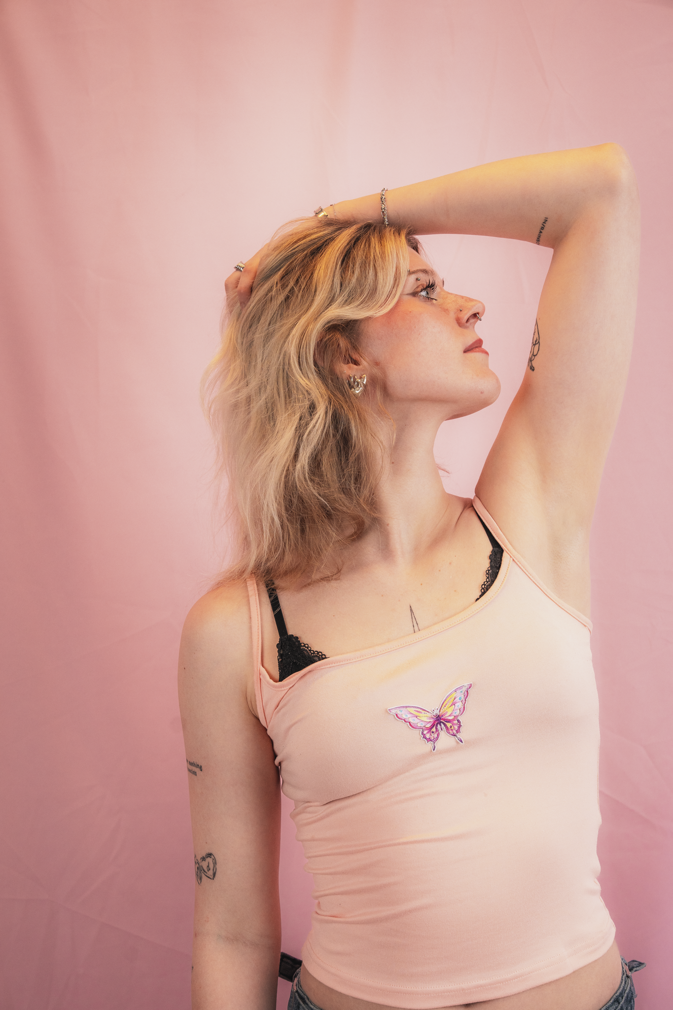 A woman with blonde hair in loose waves, wearing a peach-colored tank top with a butterfly design and black lace trim, and denim jeans, stands against a pink background. She has multiple tattoos, jewelry including earrings, rings, and a bracelet, and