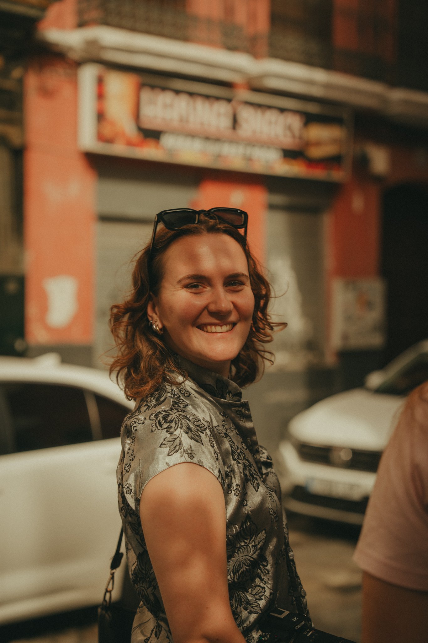 A woman with curly hair and sunglasses on her head smiling in a parking lot with cars and a sign in the background.