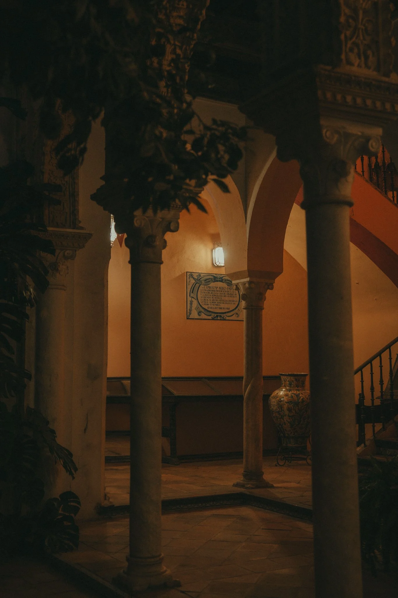 Dimly lit interior of a building with classical columns, ornate details, and a decorative vase, illuminated by warm lighting.