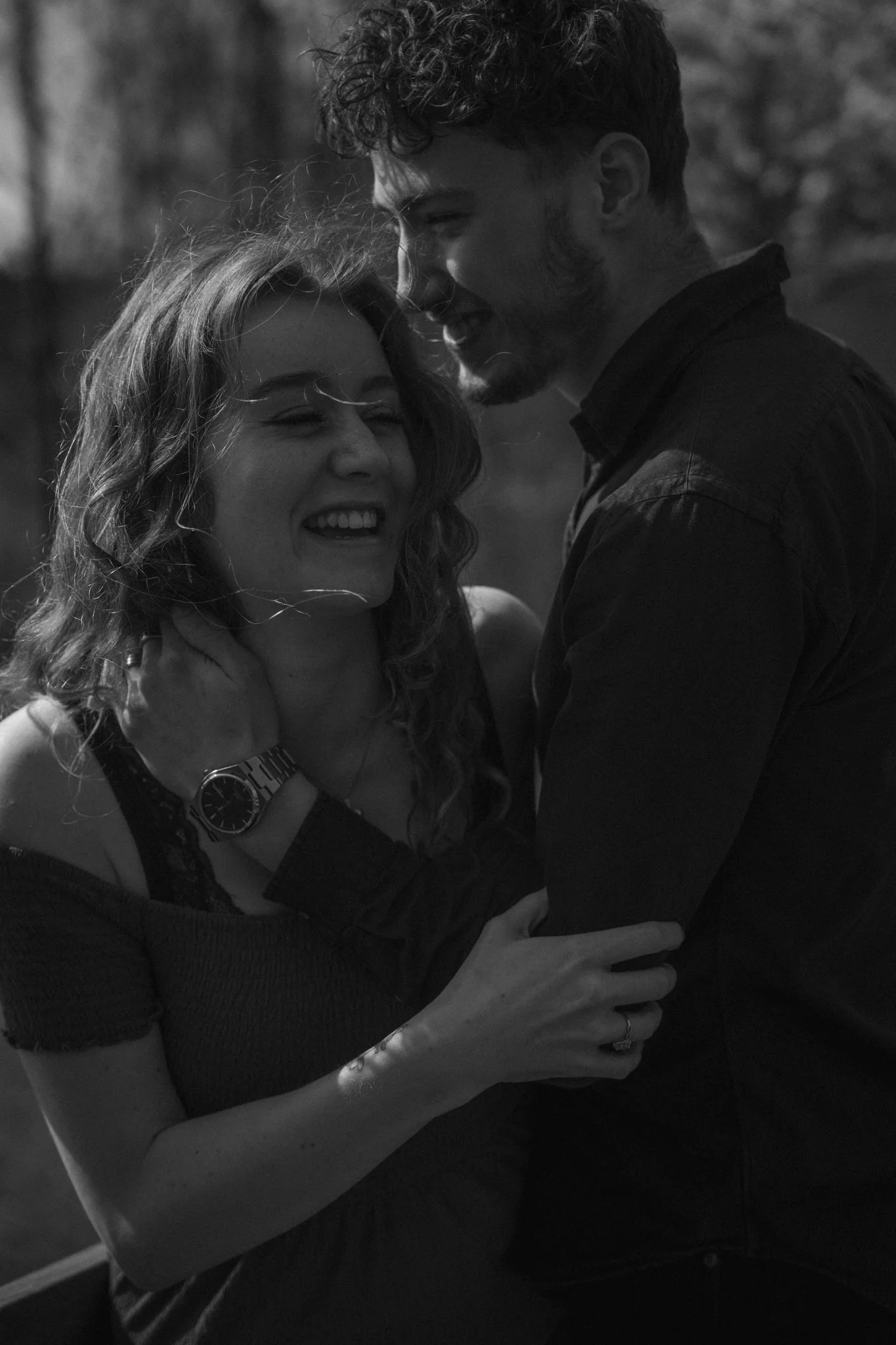 A black-and-white photo of a couple embracing outdoors, smiling, with the woman leaning into the man. The woman has curly hair, and the man has short hair and a beard.
