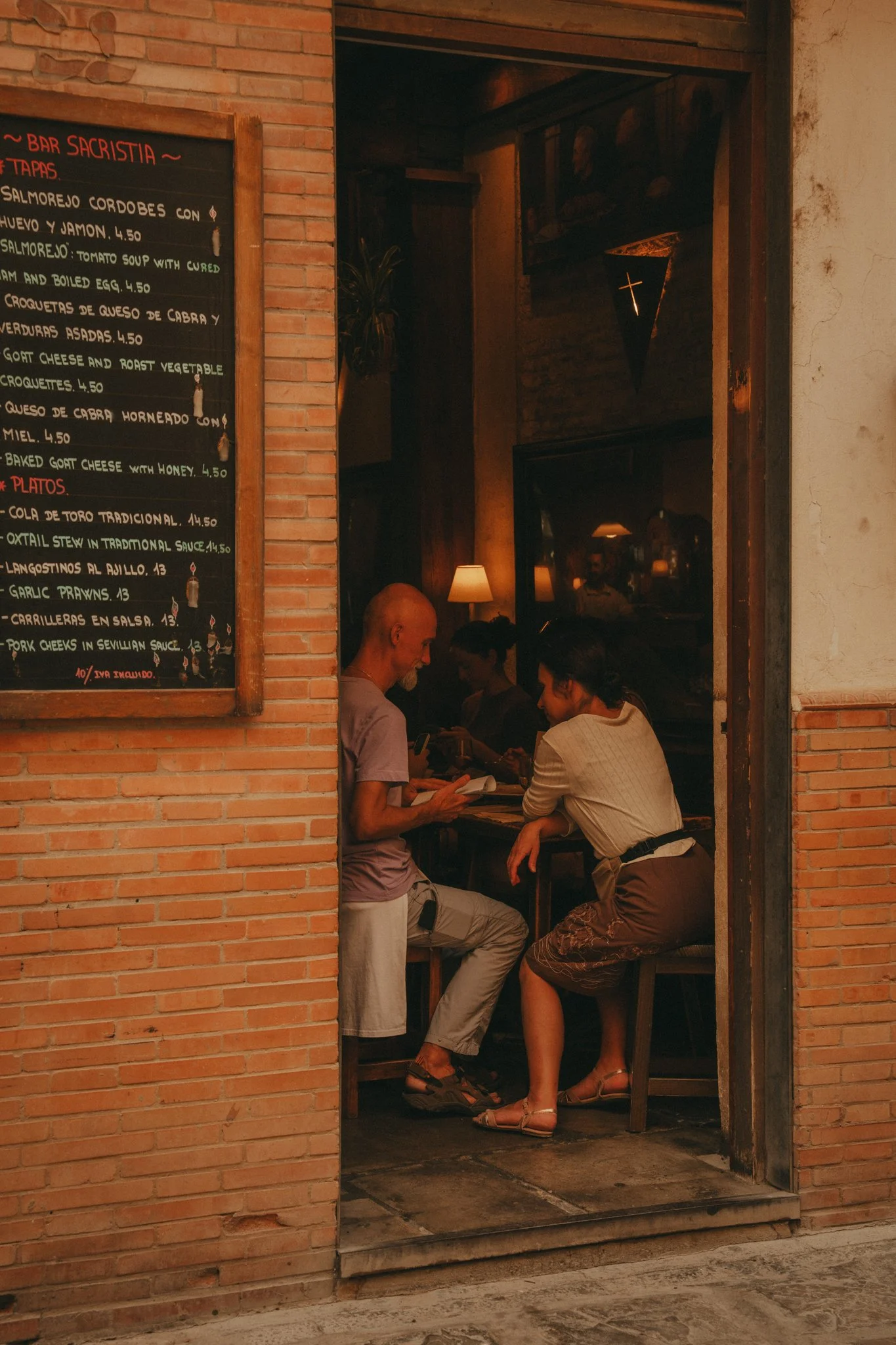 View of the interior of a restaurant with brick walls and a chalkboard menu on the left. Inside, several people are seated at a wooden counter or bar, engaged in conversation or looking at menus. Decorative lighting and a small cross sign are visible