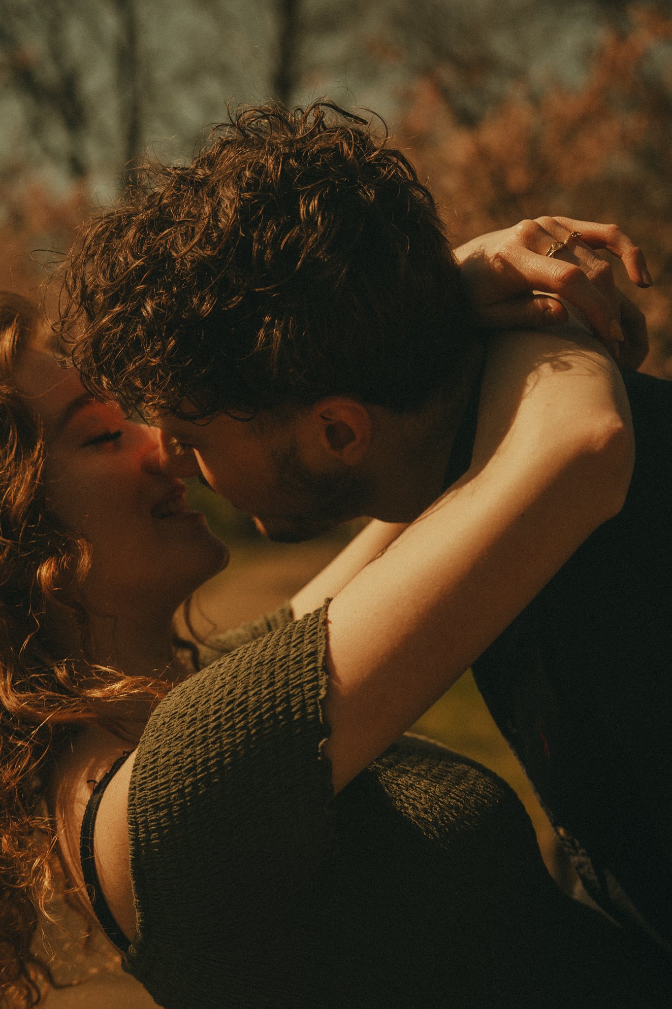 A close-up of two people with curly hair, embracing each other outdoors during sunset, with warm lighting.