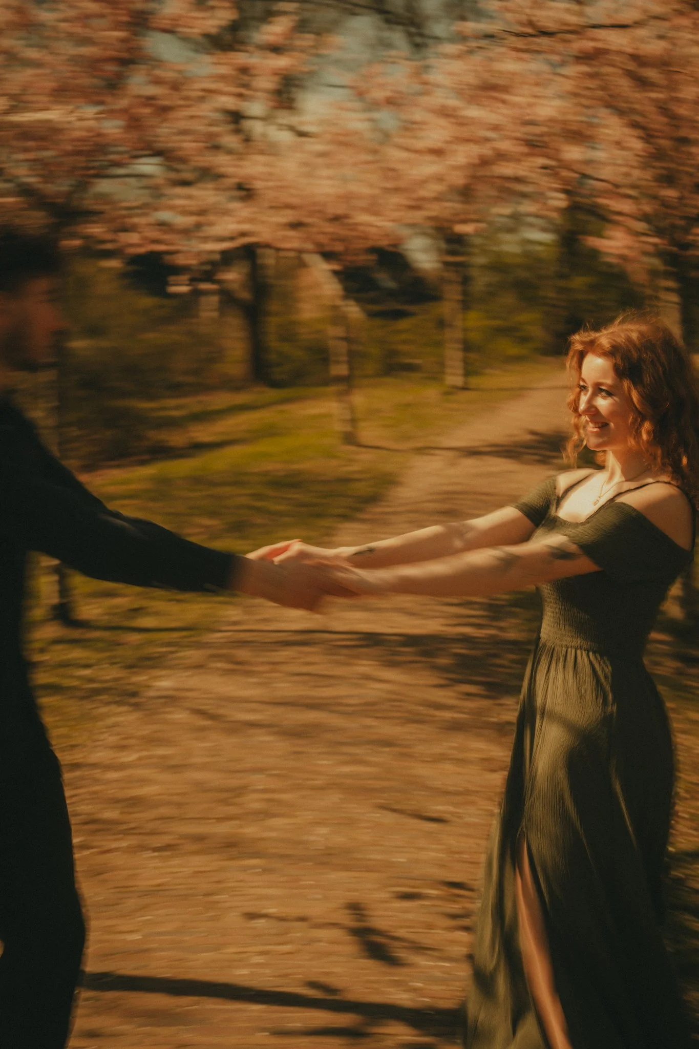 A woman in a green dress holding hands with a man in a black suit, smiling in a park with blooming cherry blossom trees.
