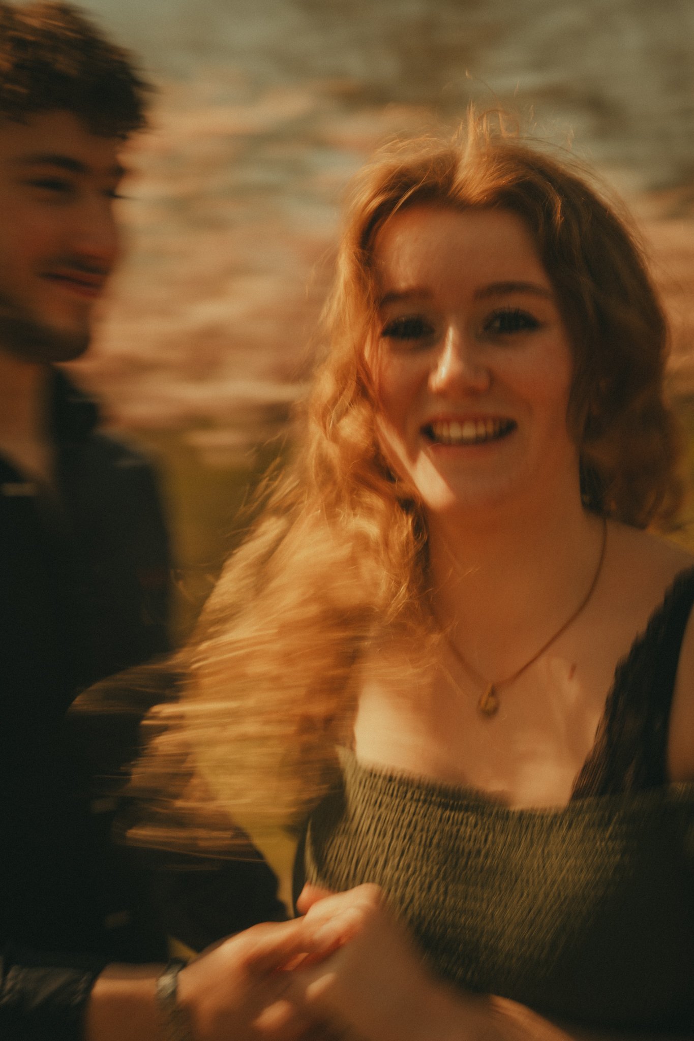 A man and a woman smiling and holding hands at night. The woman has long wavy red hair and is wearing a strapless dress and a necklace. The man is wearing a dark shirt.