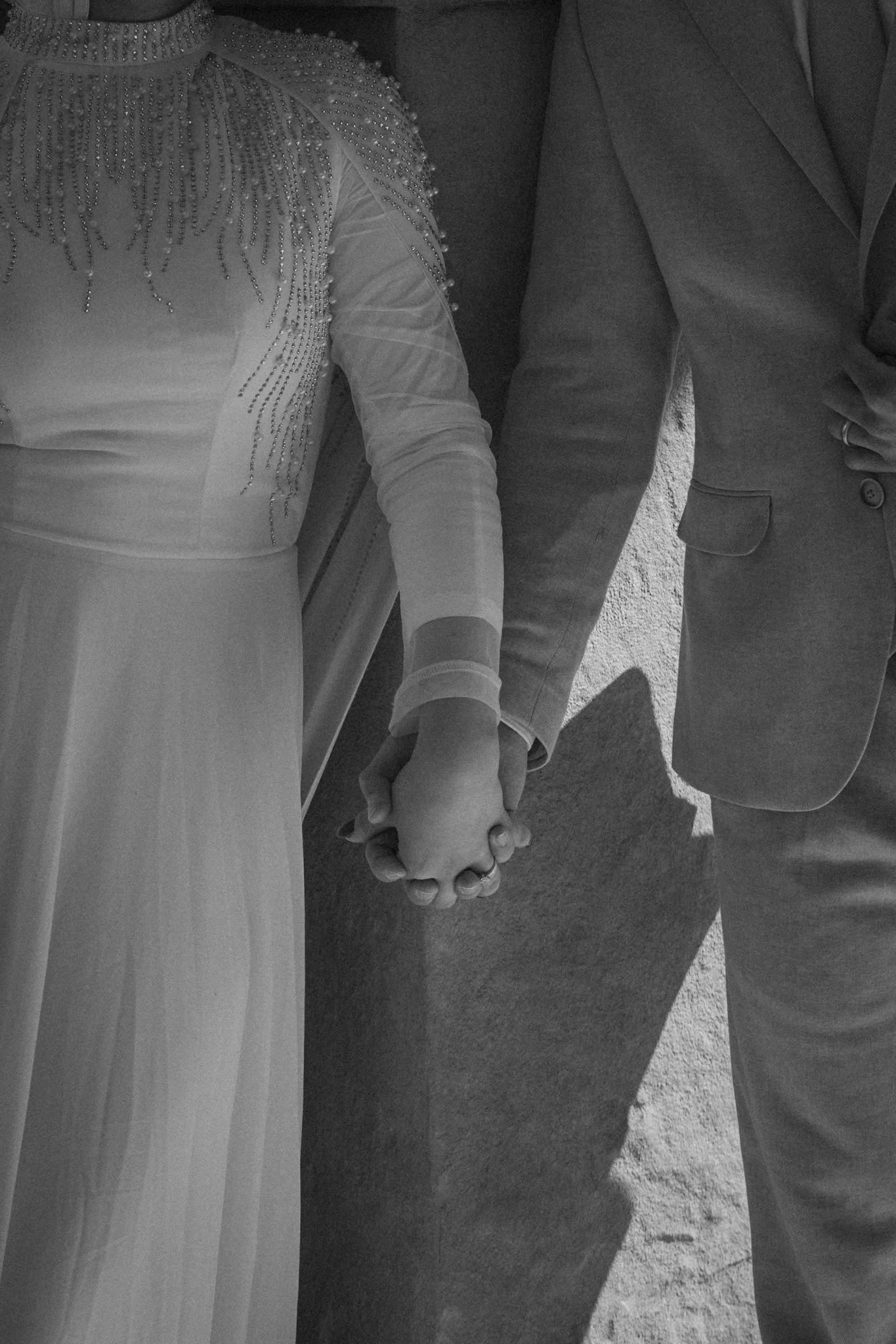 A bride and groom holding hands, wearing wedding attire, standing outdoors in sunlight.
