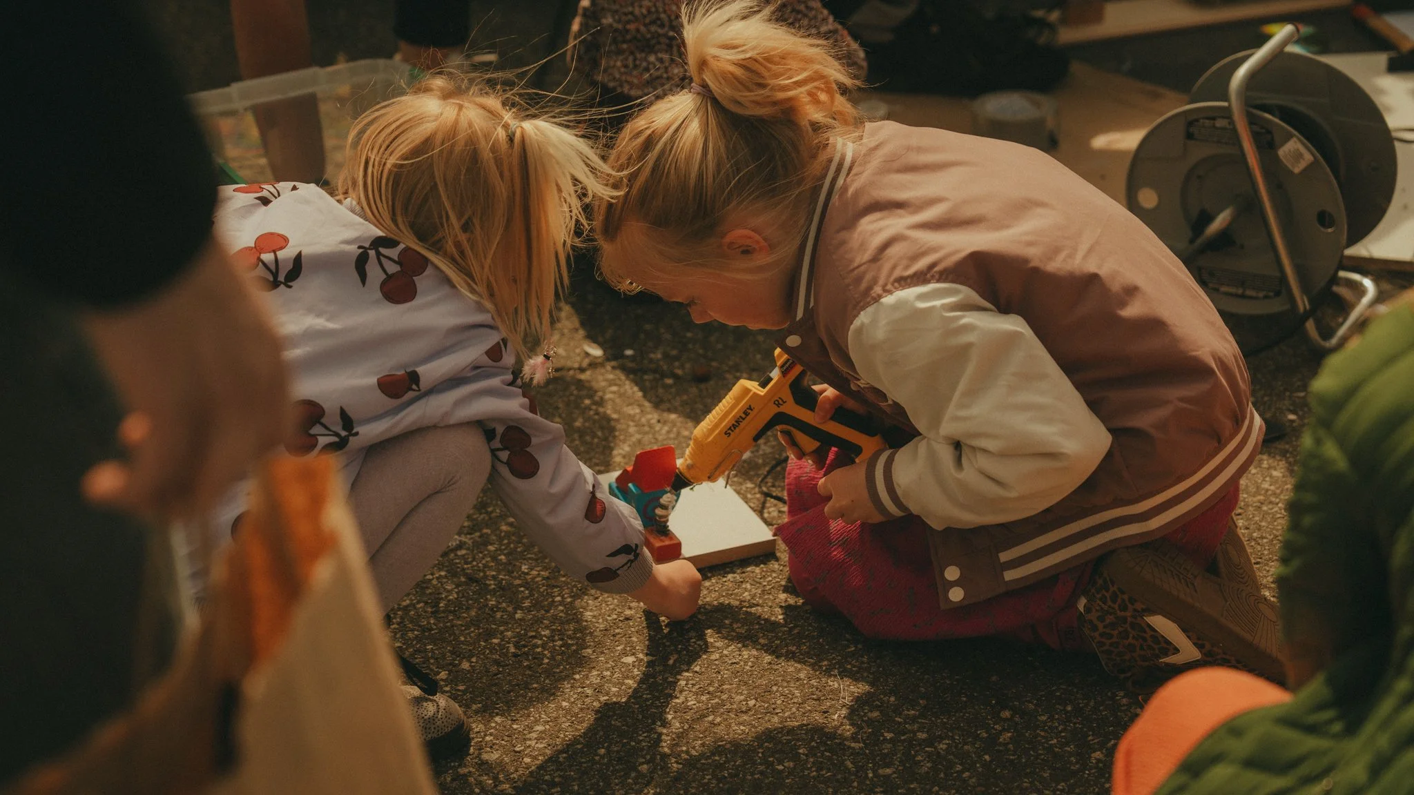 Two children kneeling on the ground and working together with a power drill, with one of them wearing a pink jacket and the other wearing a shirt with a cherry pattern.
