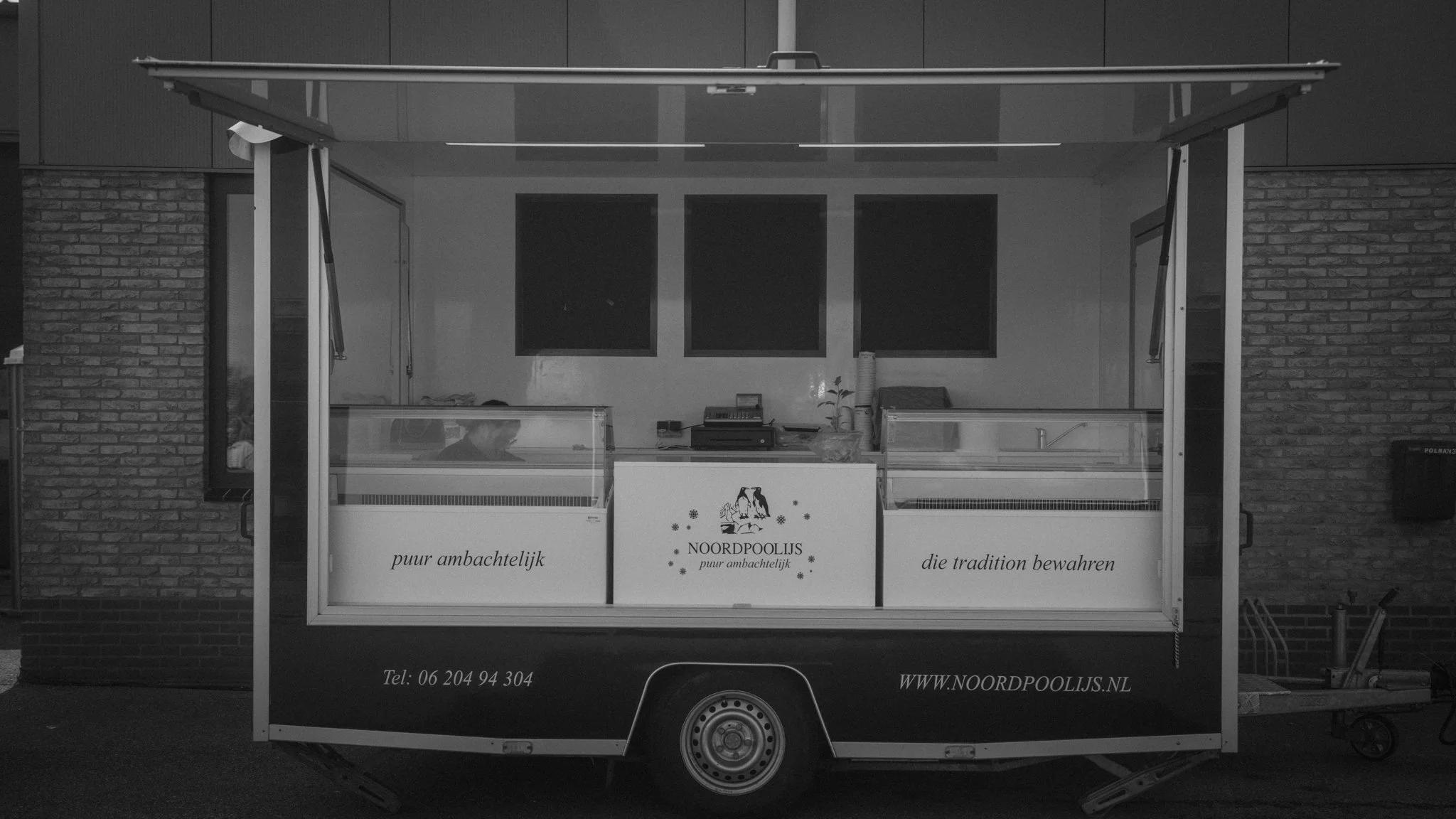 A black and white photo of a food cart with a sign that reads "NOORDPOOLIS puur ambachtelijk" and a penguin logo. The cart has a glass display case and is parked against a brick wall. There are two people inside the cart, possibly preparing or servin